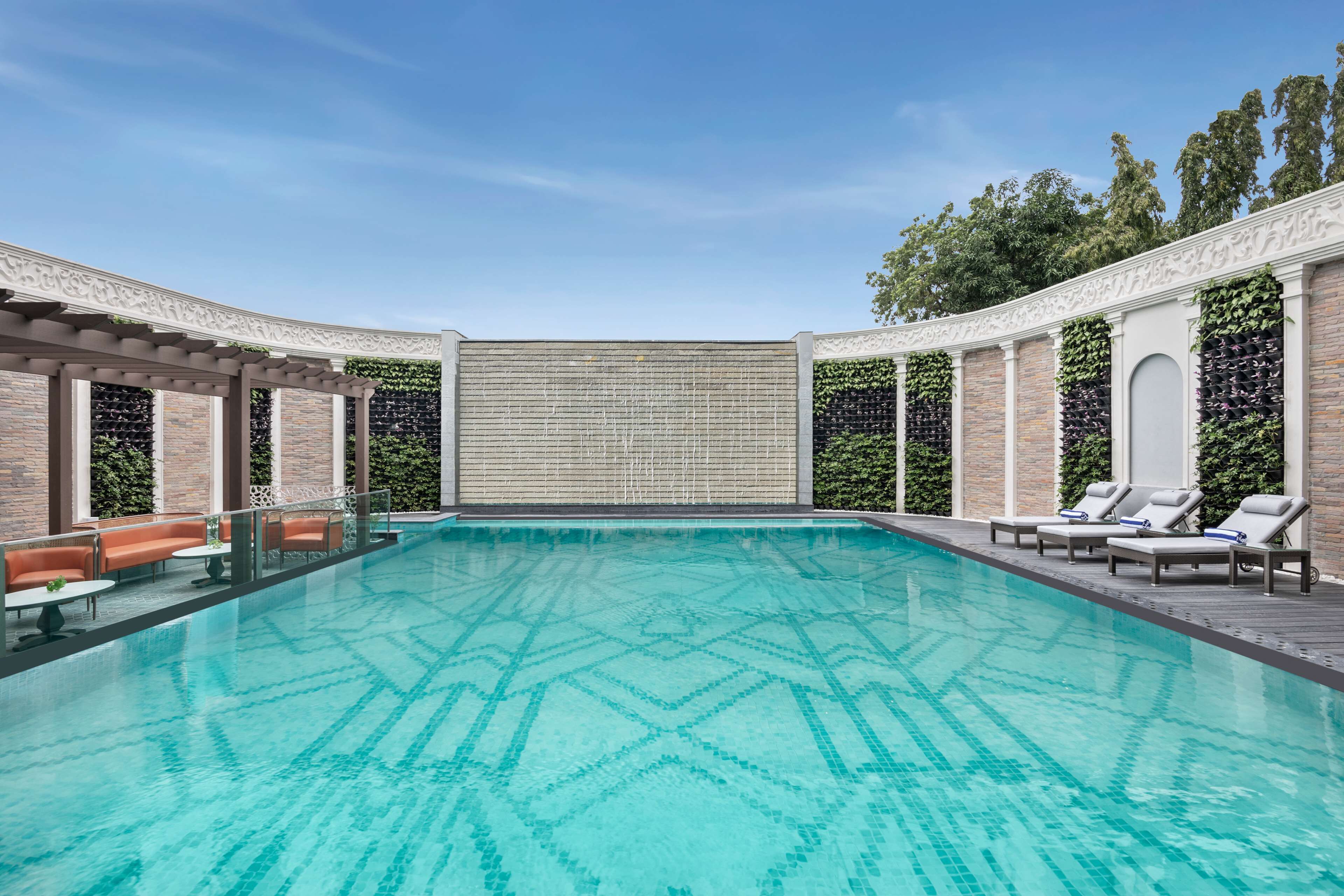 Una piscina rectangular con un diseño geométrico de azulejos, rodeada de sillas de descanso y una pérgola con pared de ladrillo y follaje.