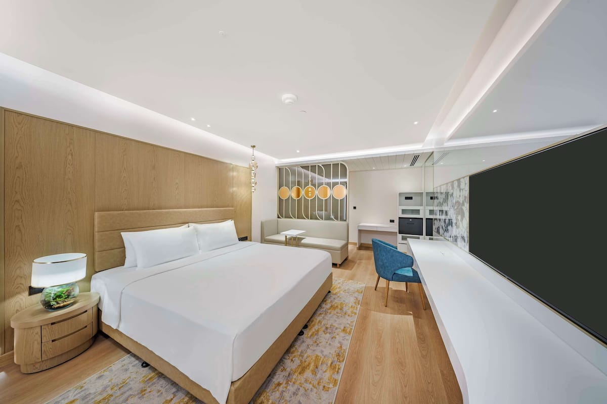 A modern hotel room with a large bed, bedside table, lamp, and a seating area with a chair and sofa.