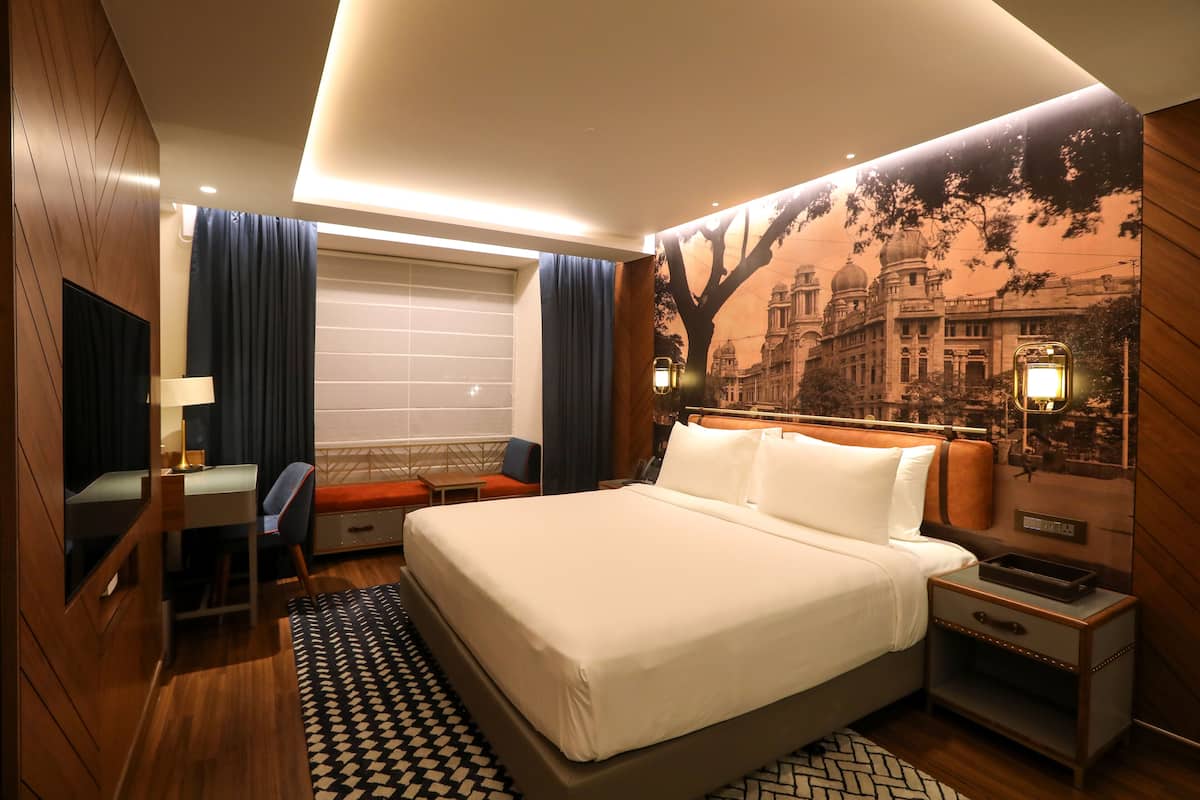A hotel room with a large bed, a desk, a chair, a nightstand, and a wall-mounted TV.