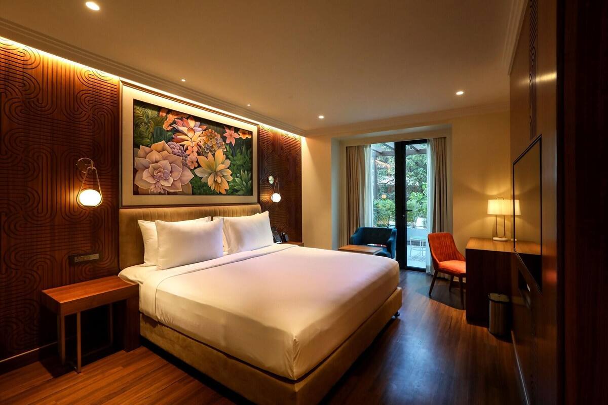 A hotel room with a large bed, a desk, a chair, a painting on the wall, and a view through the door.