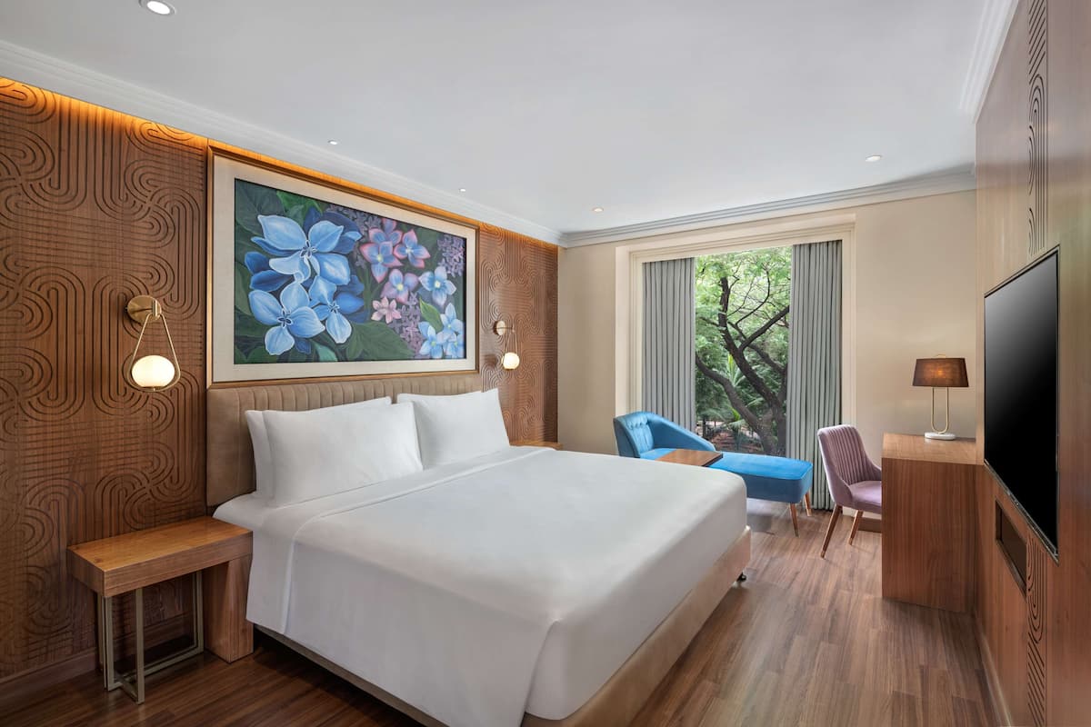 A hotel room with a large bed, a desk, a TV, and a painting of flowers on the wall.