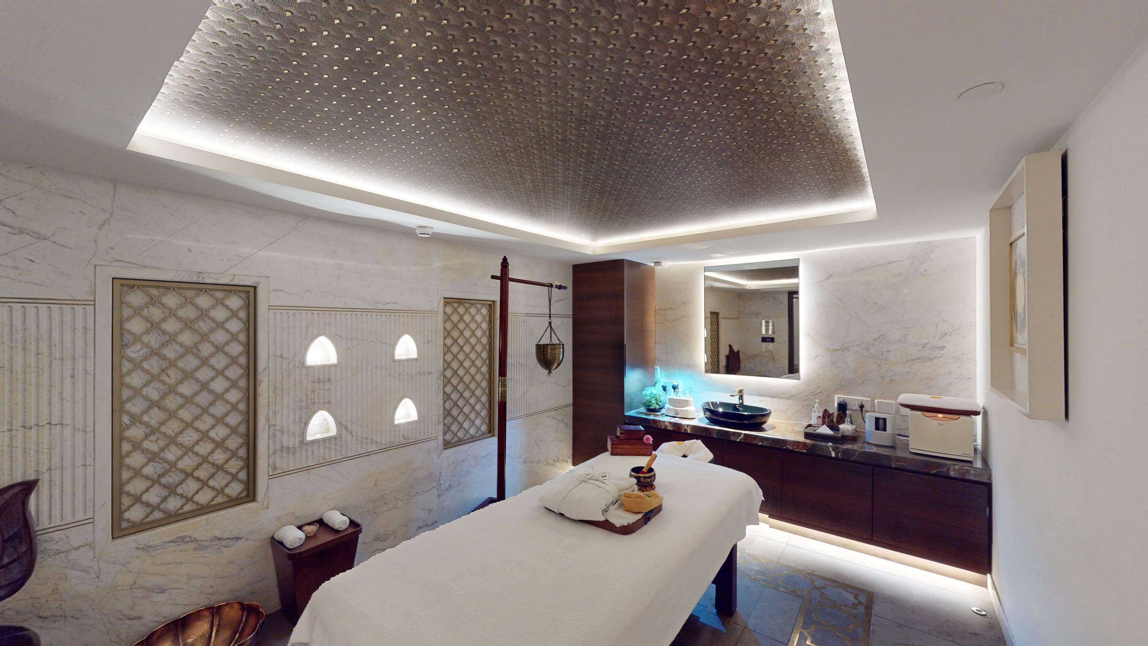 A modern spa room with a massage table, a large mirror, and a well-lit ceiling.