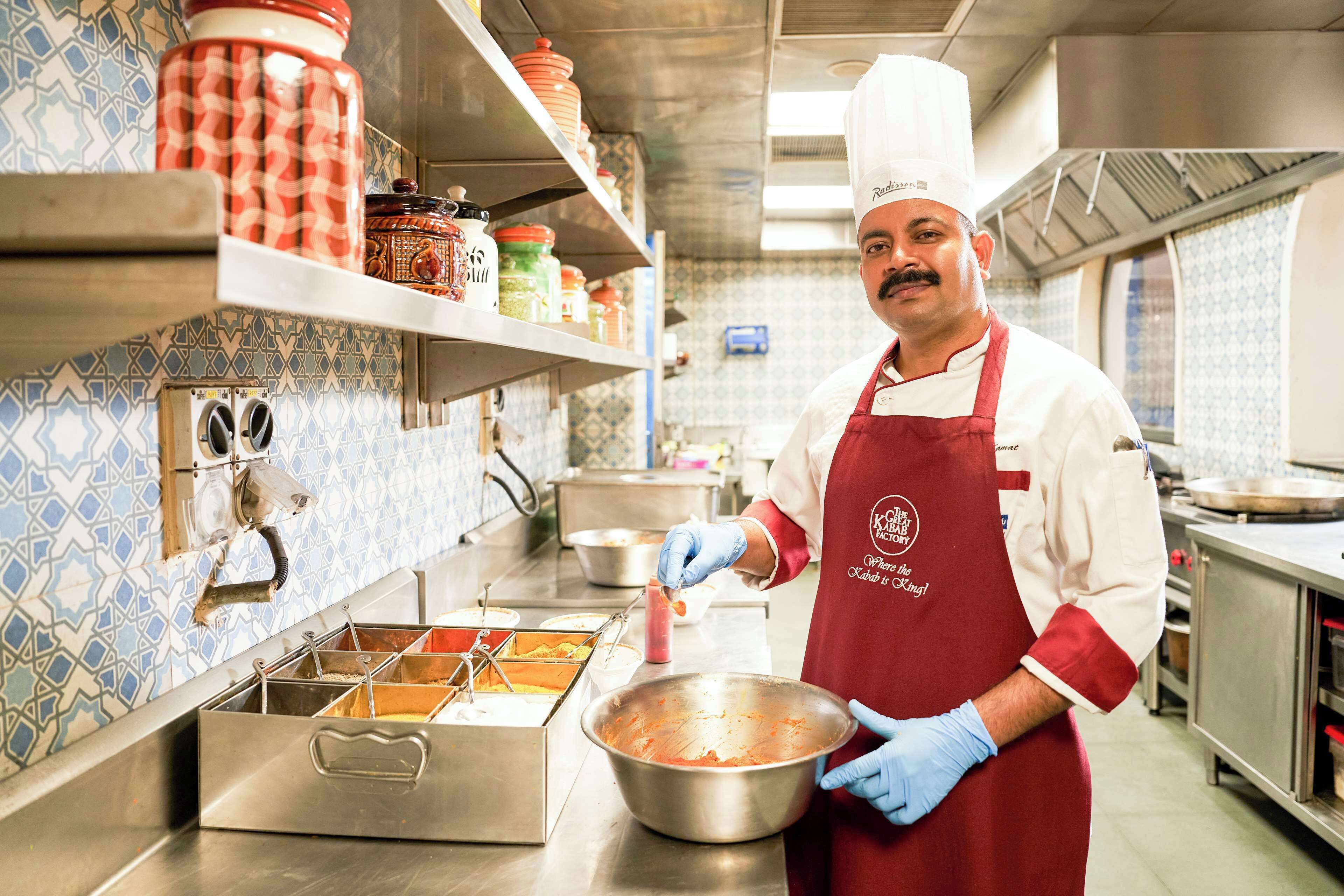 Um chef em uma cozinha com eletrodomésticos de aço inoxidável, painel de azulejos e prateleiras com potes e recipientes.