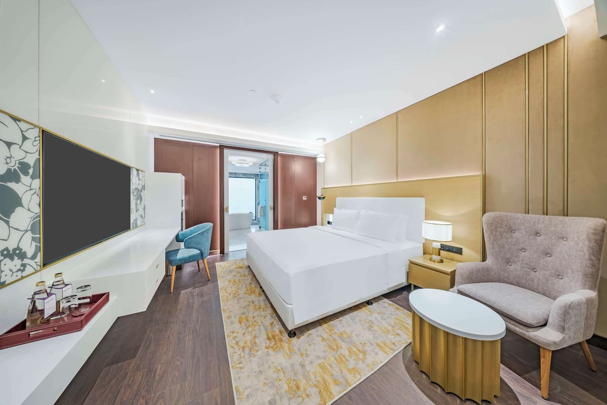 A modern hotel room with a large bed, a seating area with a chair, a small table, and a bedside lamp.