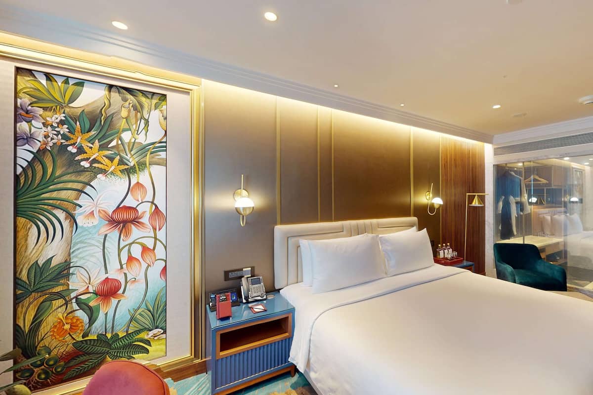 A hotel room with a large bed, a nightstand with a phone and a lamp, a wall with a floral painting, and a sliding door leading to a bathroom.