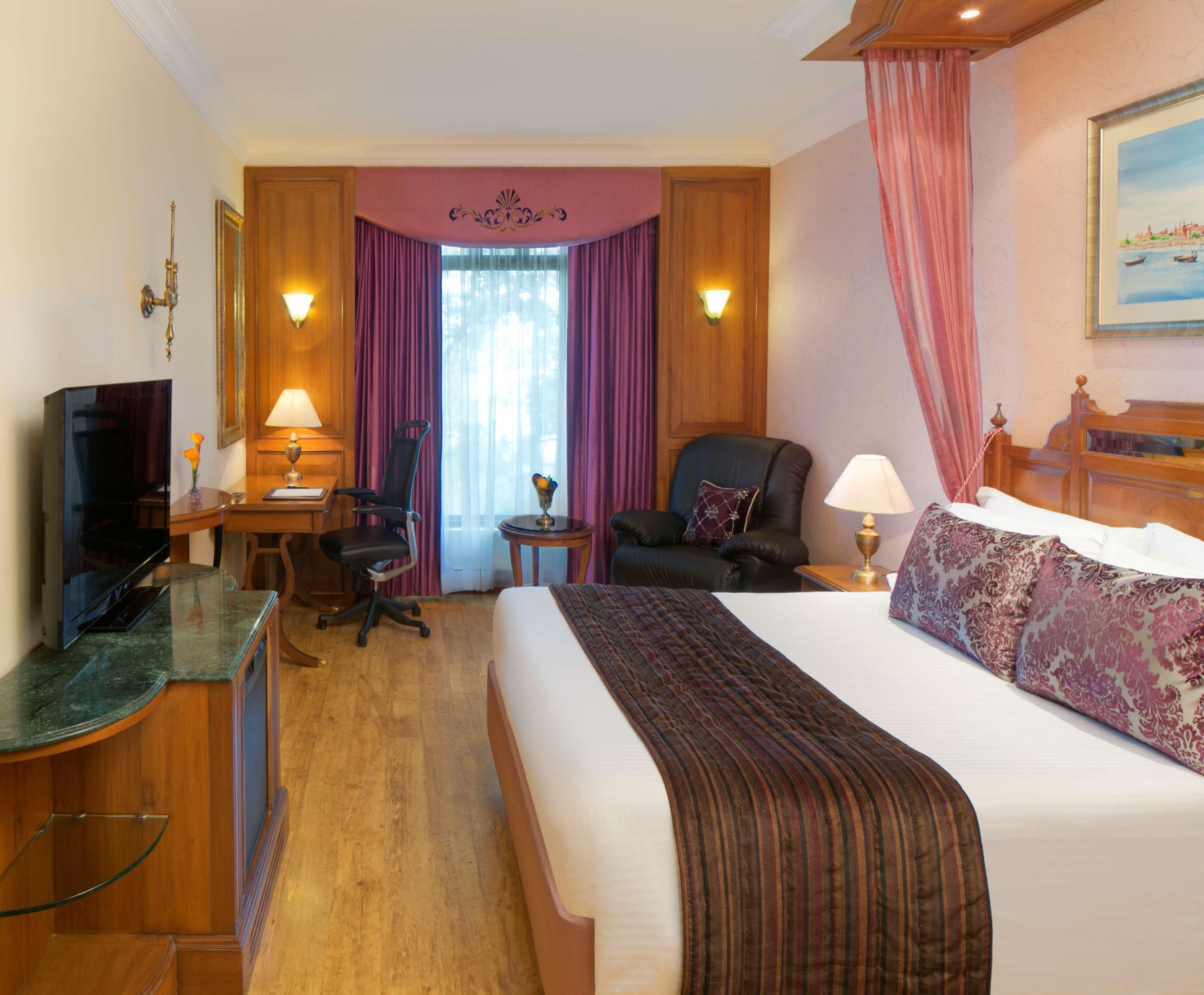 Habitación de hotel con cama, escritorio con televisor, silla, lámpara y ventana con cortinas.