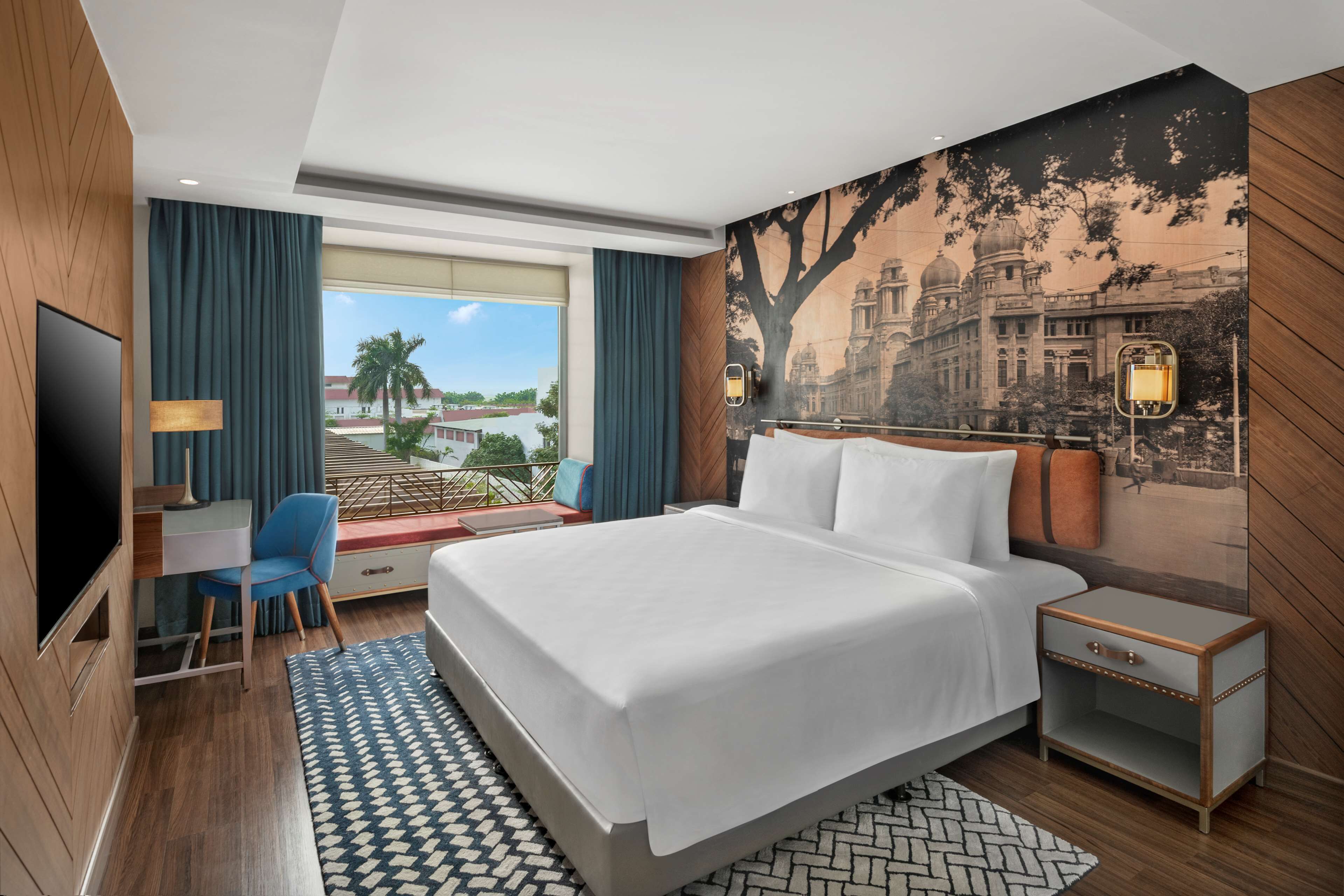 A hotel room with a large bed, a desk, a chair, a TV, and a view of a cityscape.