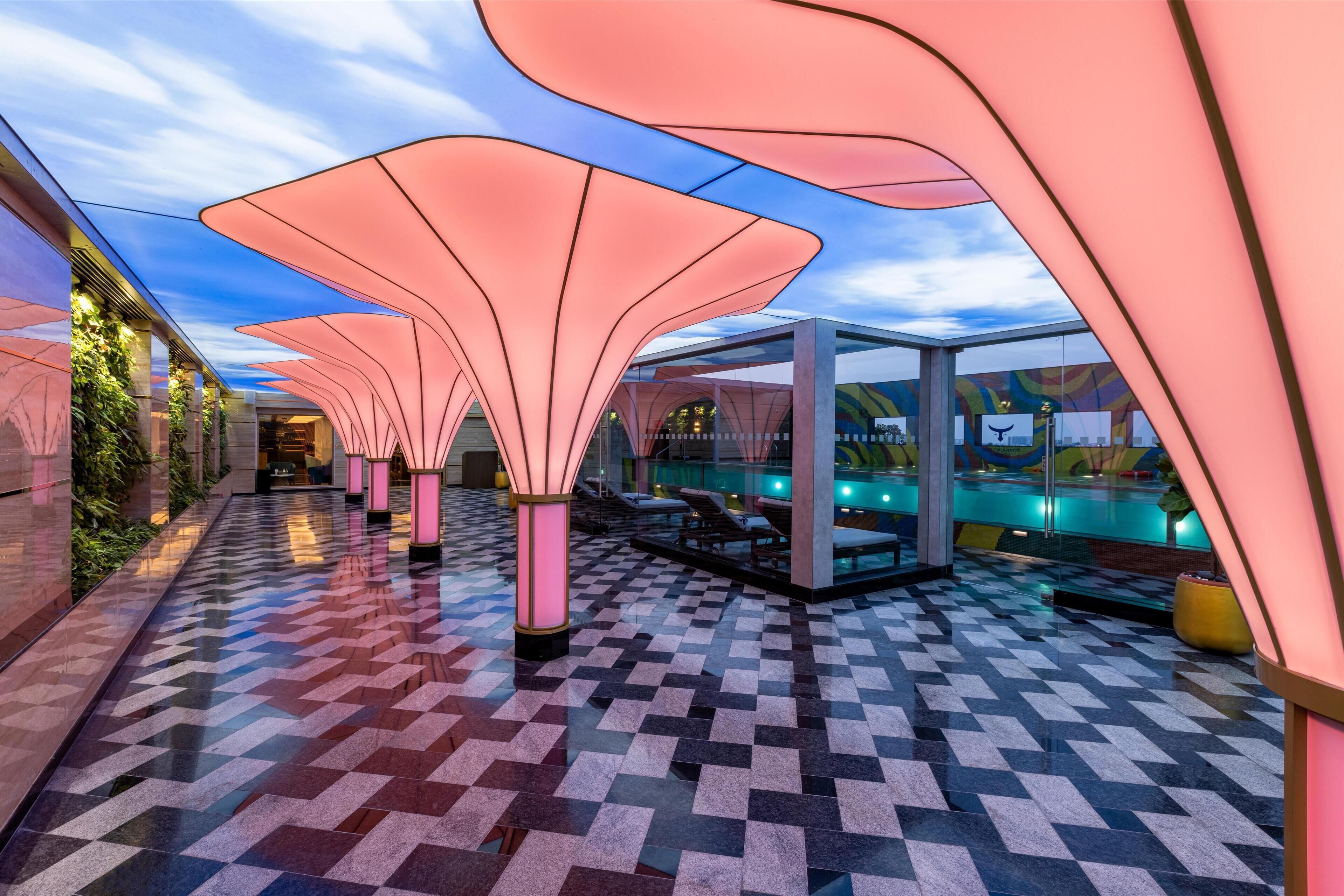 Un pasillo moderno de hotel con suelo estampado, grandes estructuras iluminadas y una zona de piscina con sillones de descanso.