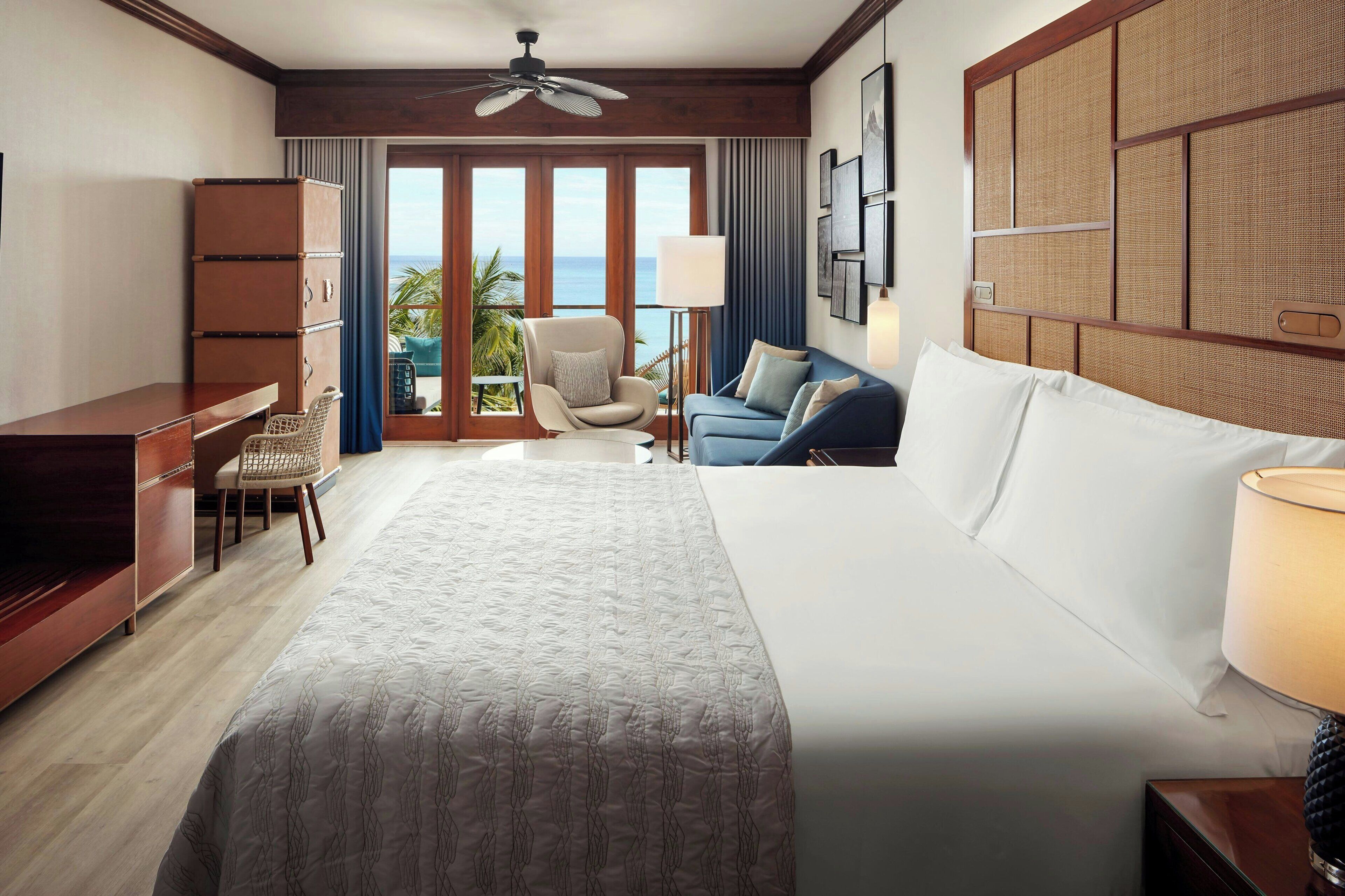 A hotel room with a bed, a desk, a chair, a sofa, a lamp, and a view of the ocean.