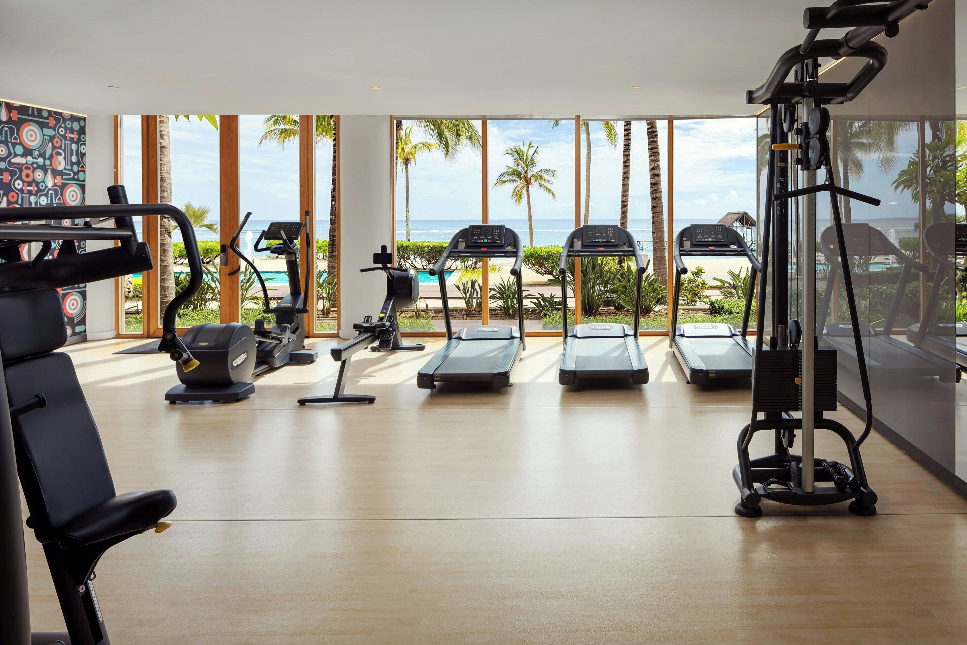Un gimnasio con cintas de correr, una máquina de remo y una bicicleta estática, con vista al océano y palmeras.
