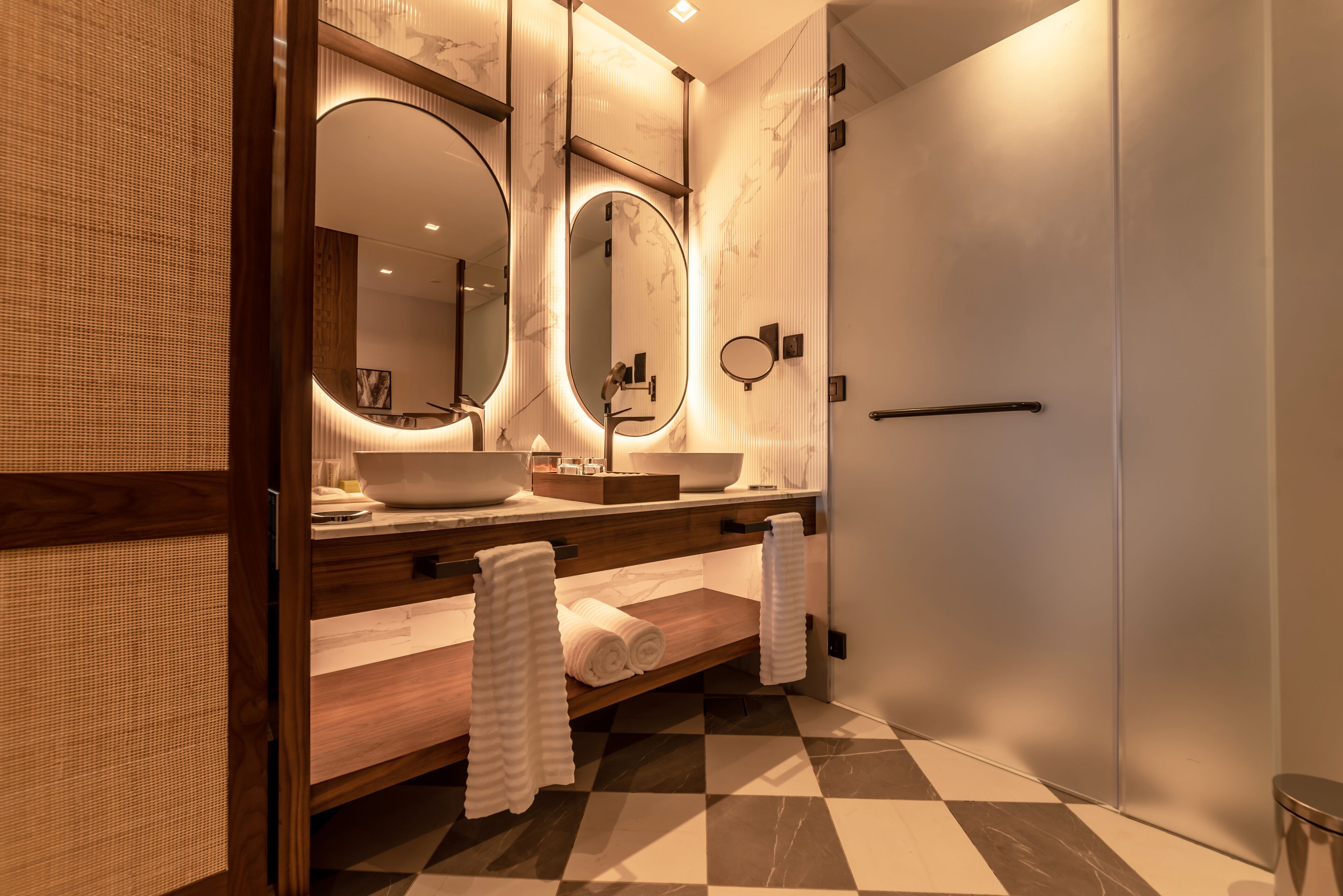 A modern bathroom with a wooden vanity, two large sinks, a mirror with integrated lighting, and a checkered floor.