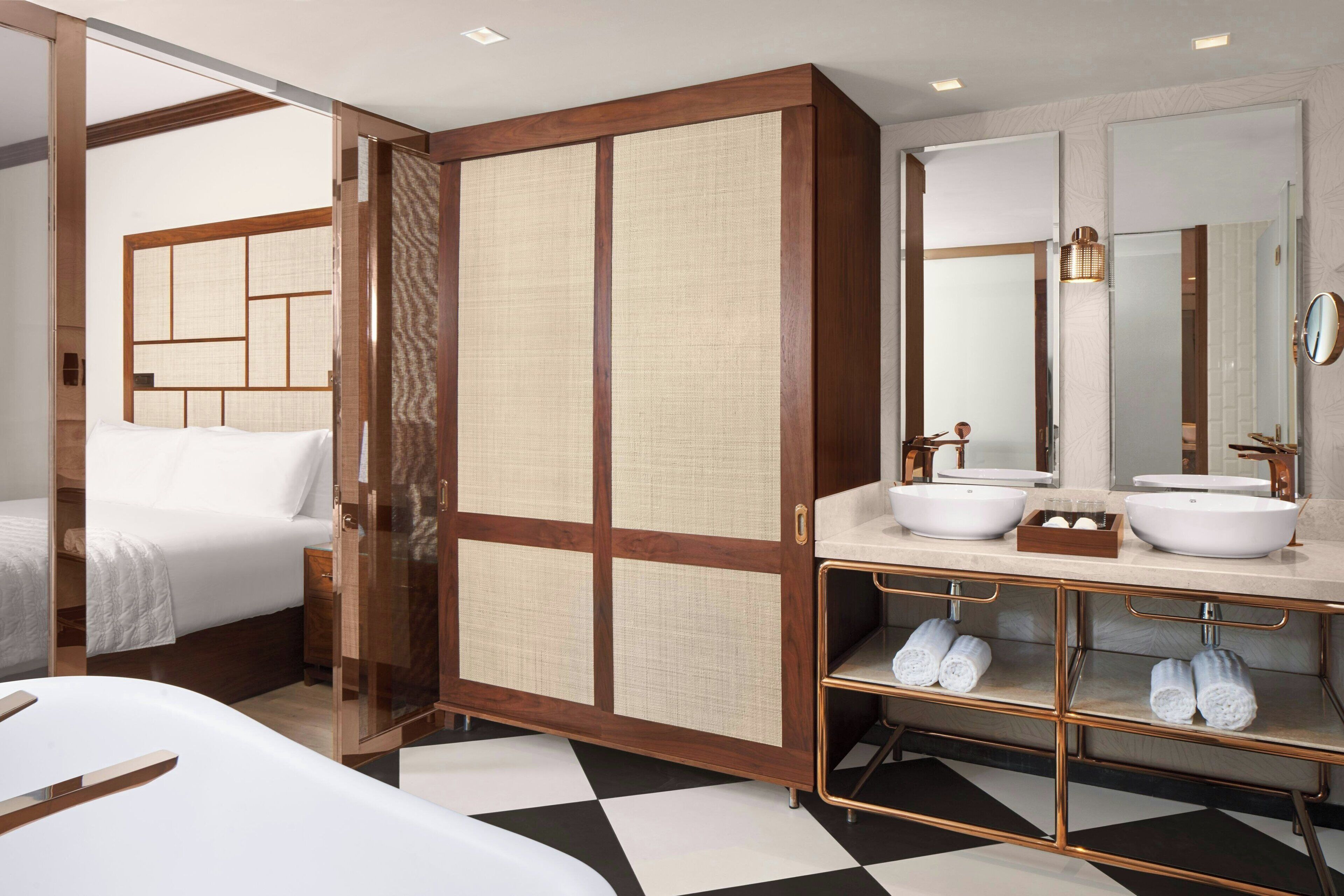 A hotel room with a bed, a wooden wardrobe, a bathroom with two sinks and a mirror, and a checkered floor.