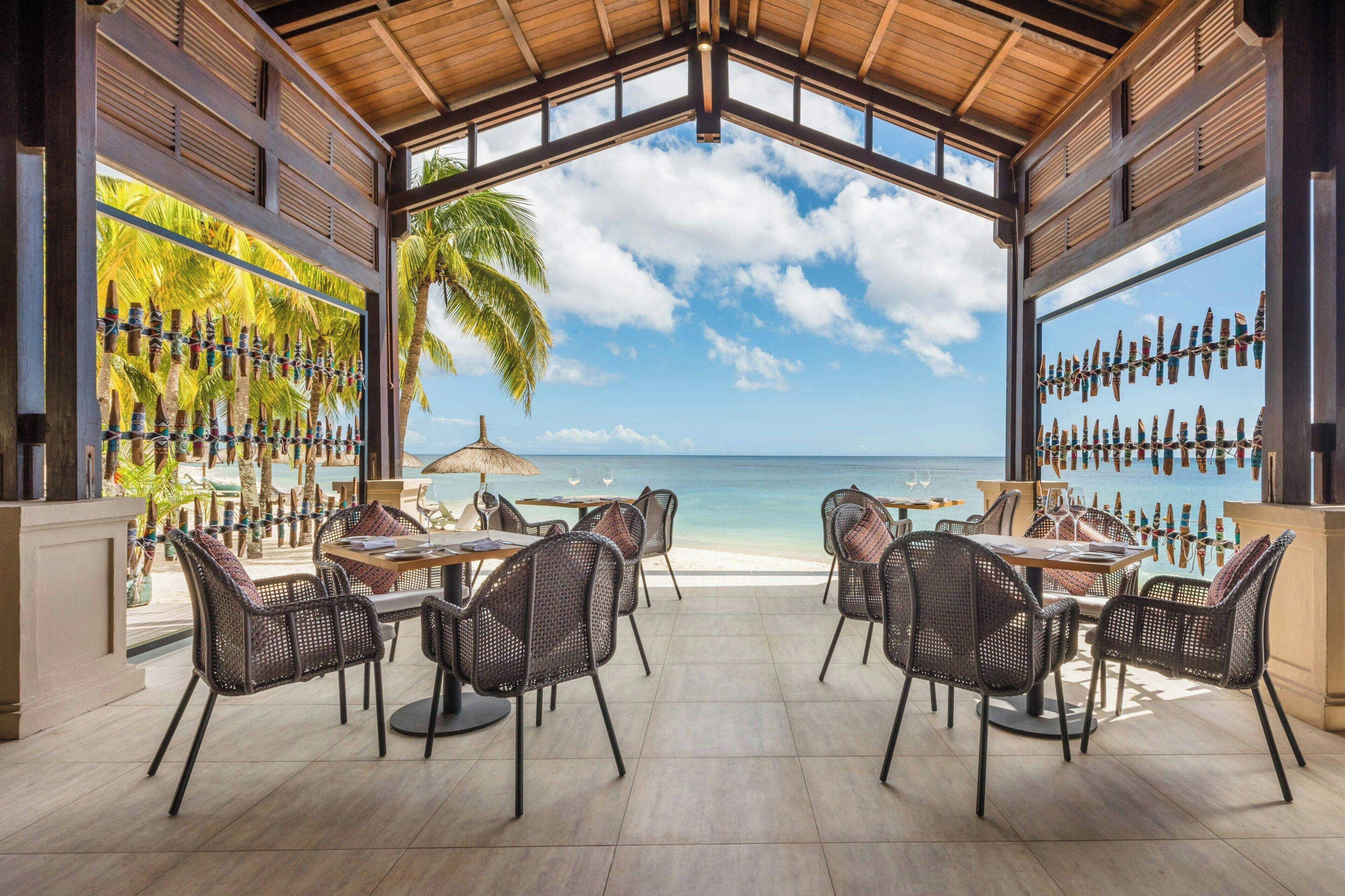 An outdoor dining area with wicker chairs and tables, overlooking a beach and ocean.