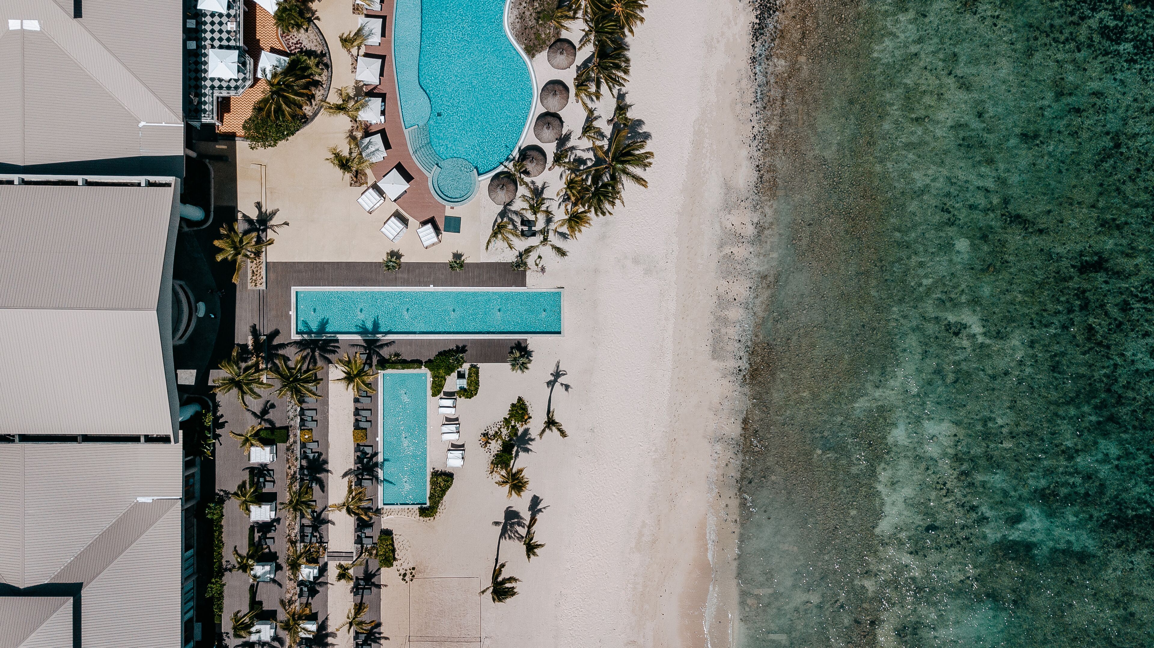 An aerial view of a resort with a large swimming pool, beach area, and lounge chairs.