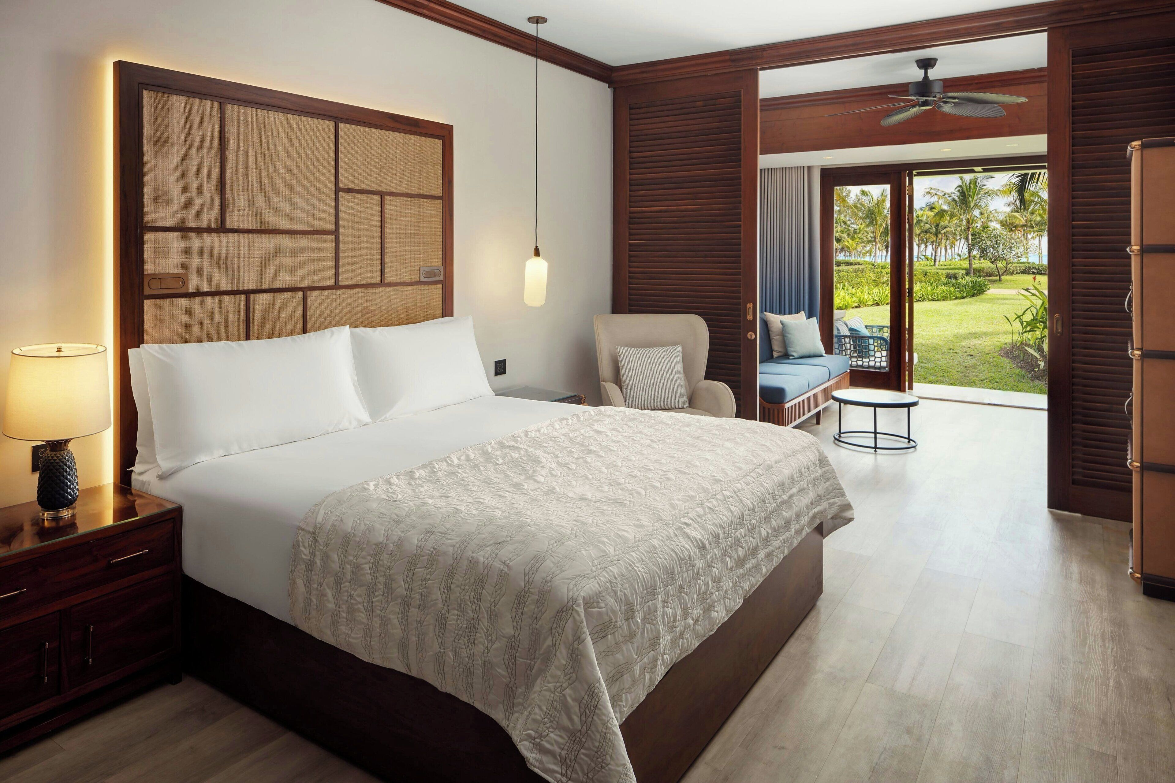 A hotel room with a large bed, a nightstand with a lamp, a chair, a small table, and a view of the outdoors.