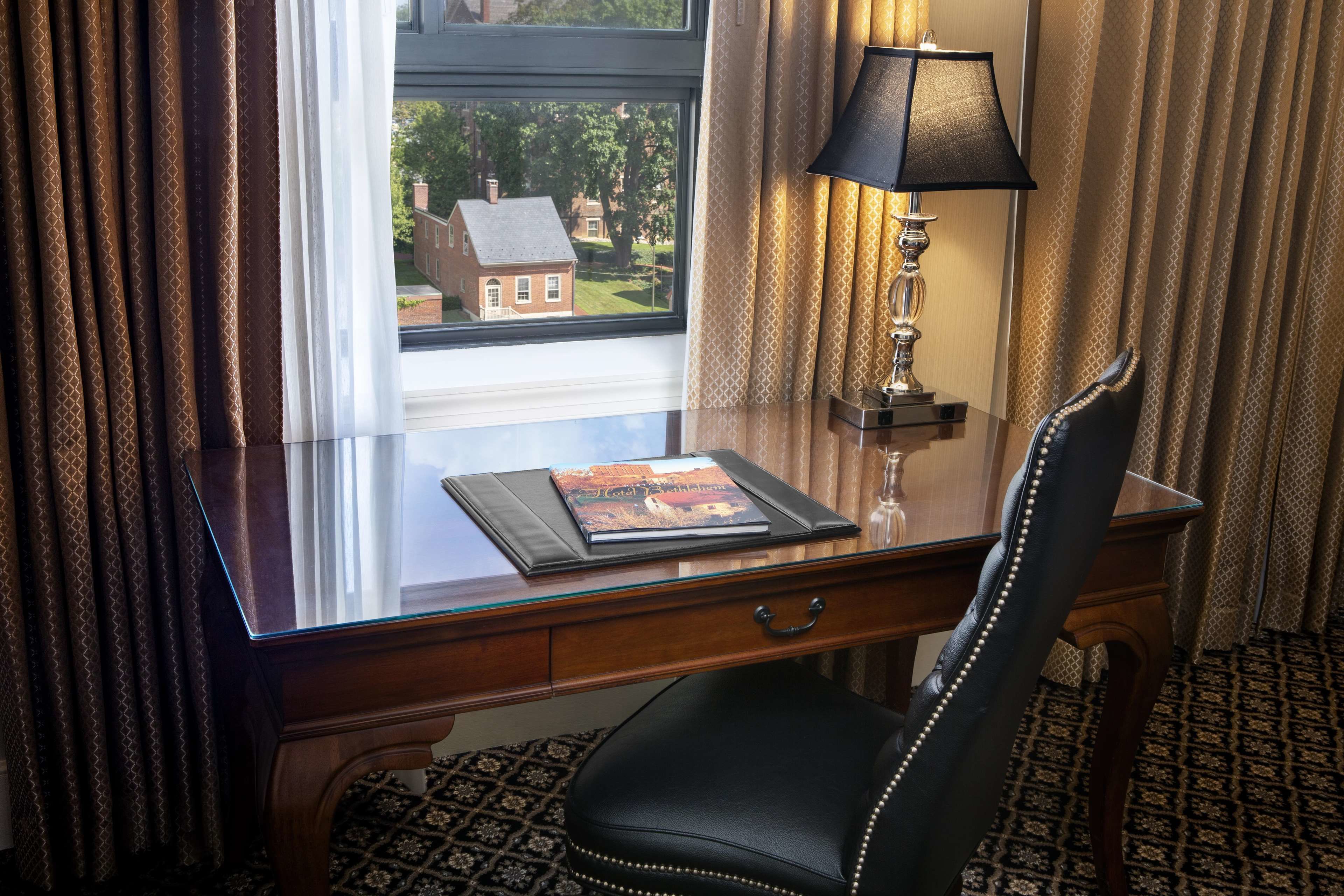 A hotel room with a wooden desk, a black leather chair, a lamp, and a view of a house through the window.