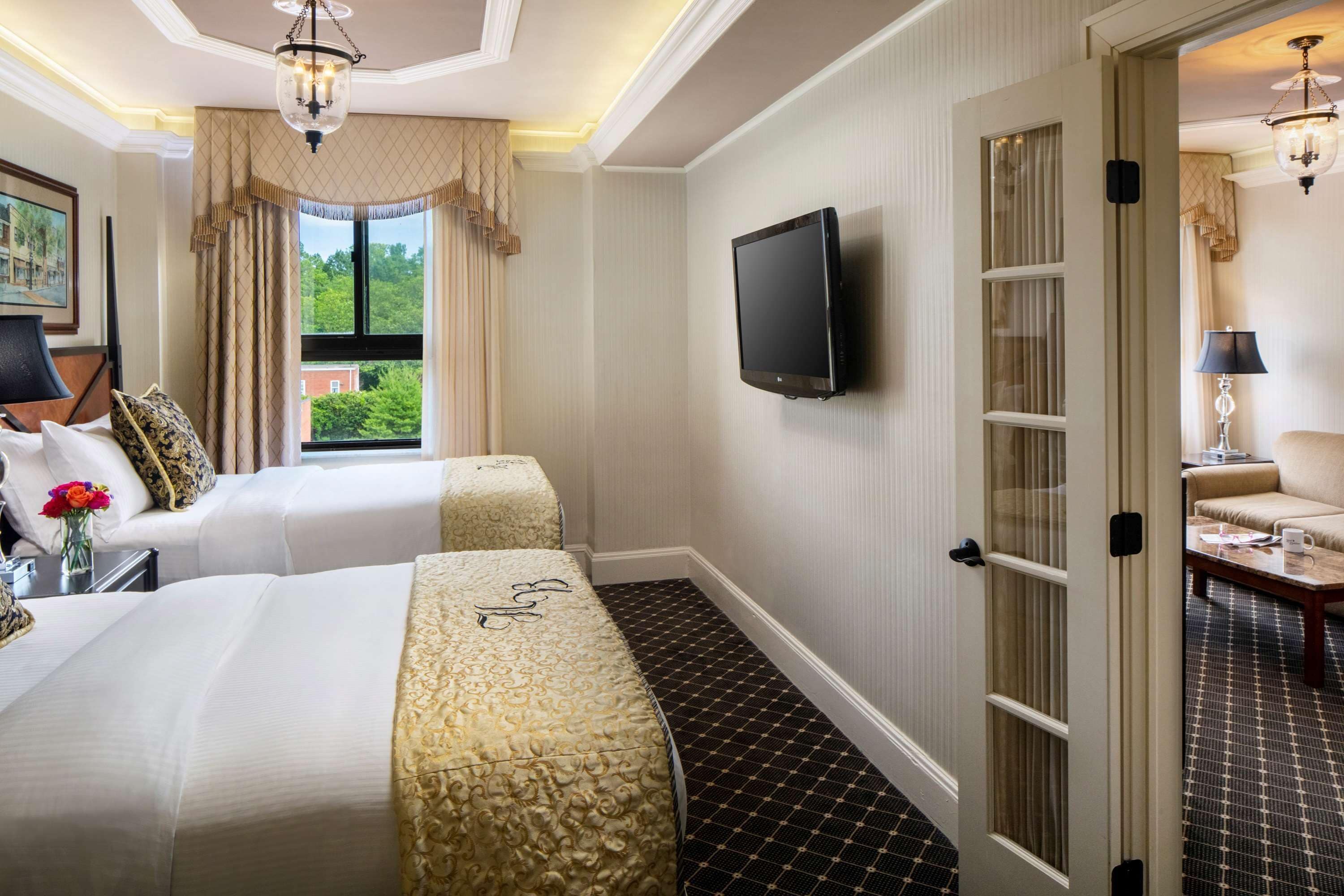 A hotel room with two beds, a TV, a window with curtains, and a view of greenery outside.