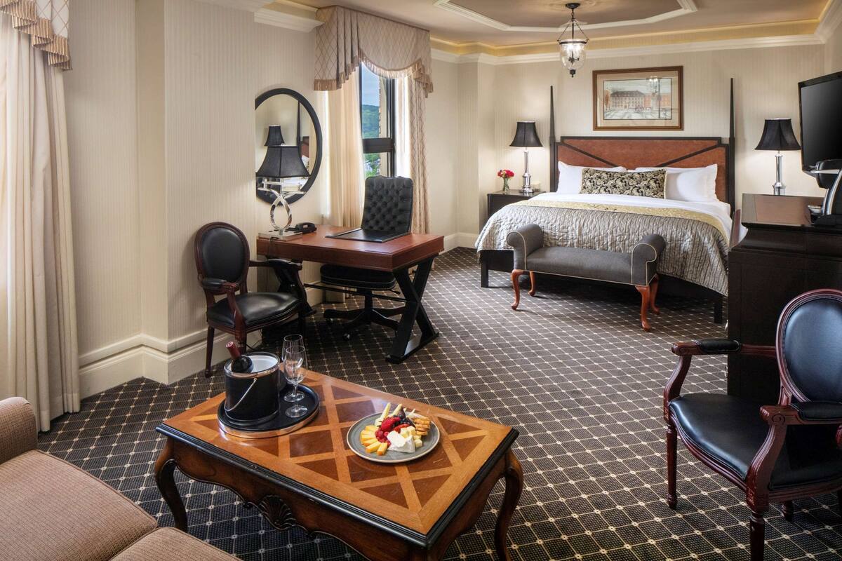 Ein Hotelzimmer mit einem Bett, einem Schreibtisch mit Stuhl, einem Tisch mit einer Speiseplatte, einem Fernseher und einem Fenster mit Vorhängen.