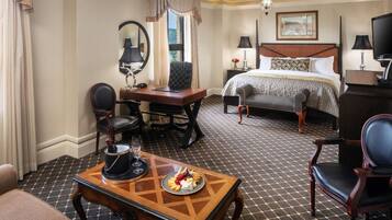 A hotel room with a bed, a desk with a chair, a table with a plate of food, a TV, and a window with curtains.
