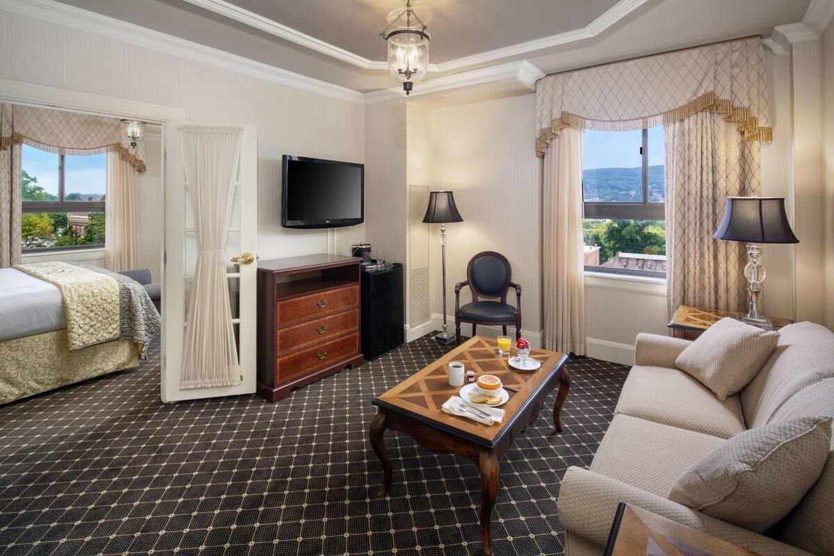 Ein Hotelzimmer mit Bett, Sofa, Fernseher, Tisch mit Frühstücksangeboten und Blick ins Freie.