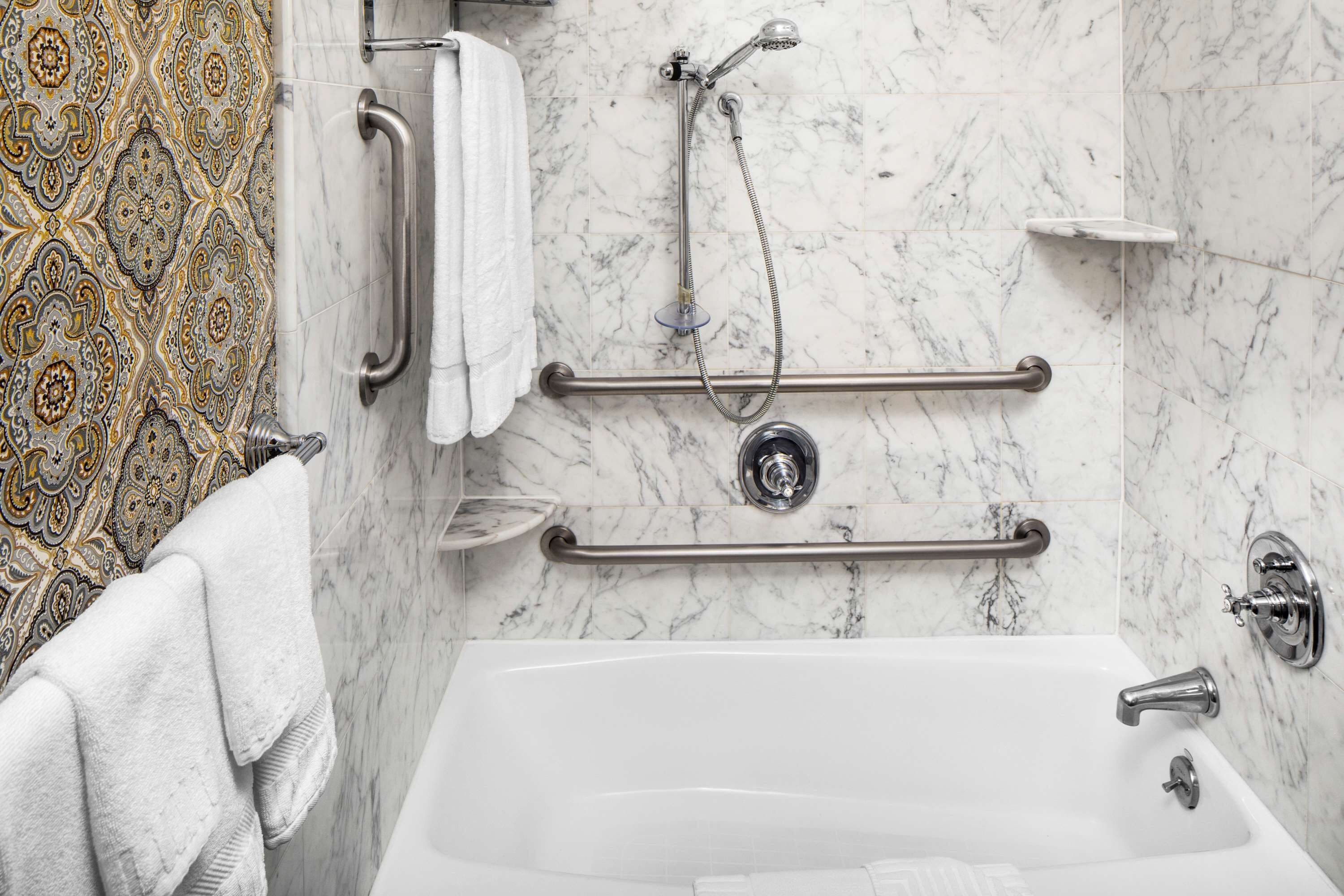 Ein Badezimmer mit einer weißen Wanne, Haltegriffen, einer Duschbrause und einer Wand mit Muster.