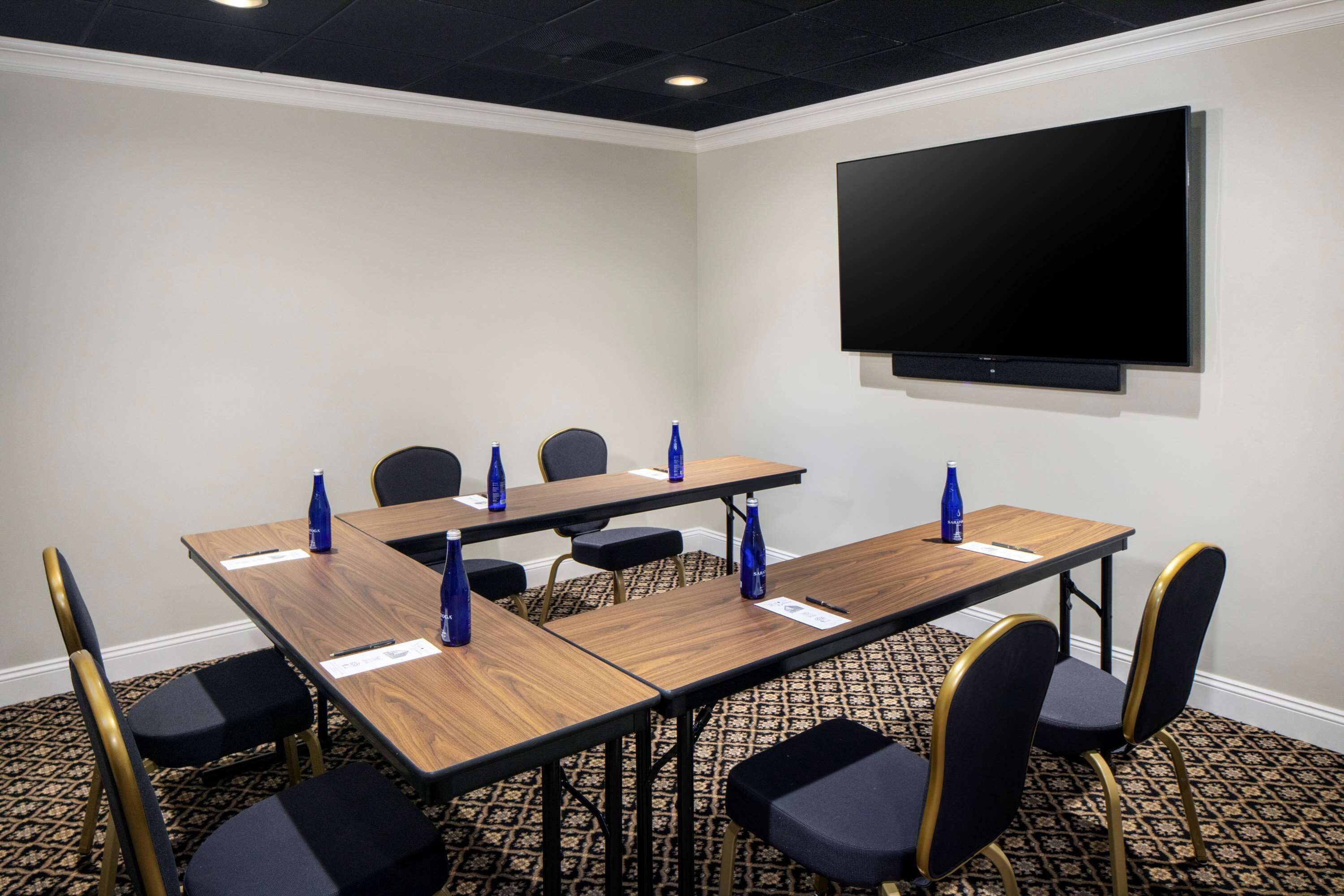 Uma sala de conferência com uma mesa comprida, cadeiras, uma TV de tela plana e garrafas de água azuis sobre a mesa.