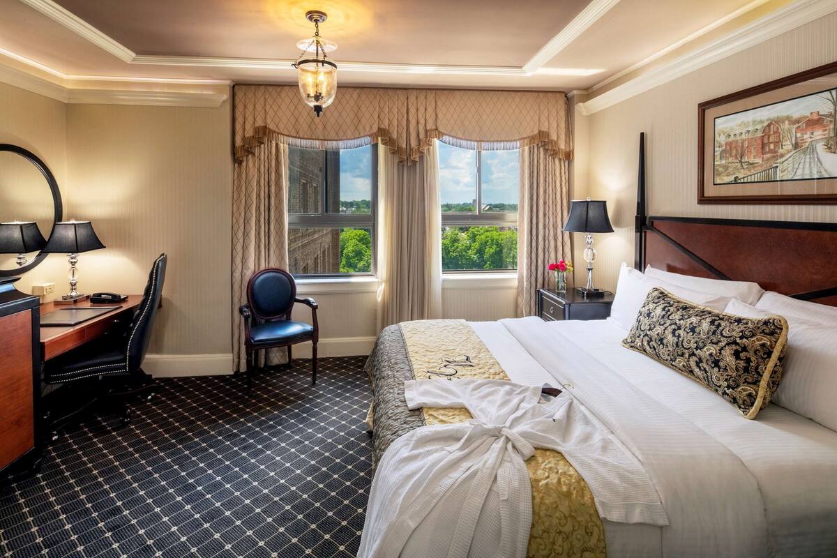 Ein Hotelzimmer mit einem großen Bett, einem Schreibtisch mit Stuhl, einer Schreibtischlampe, einem Sessel und einem Fenster mit Vorhängen.