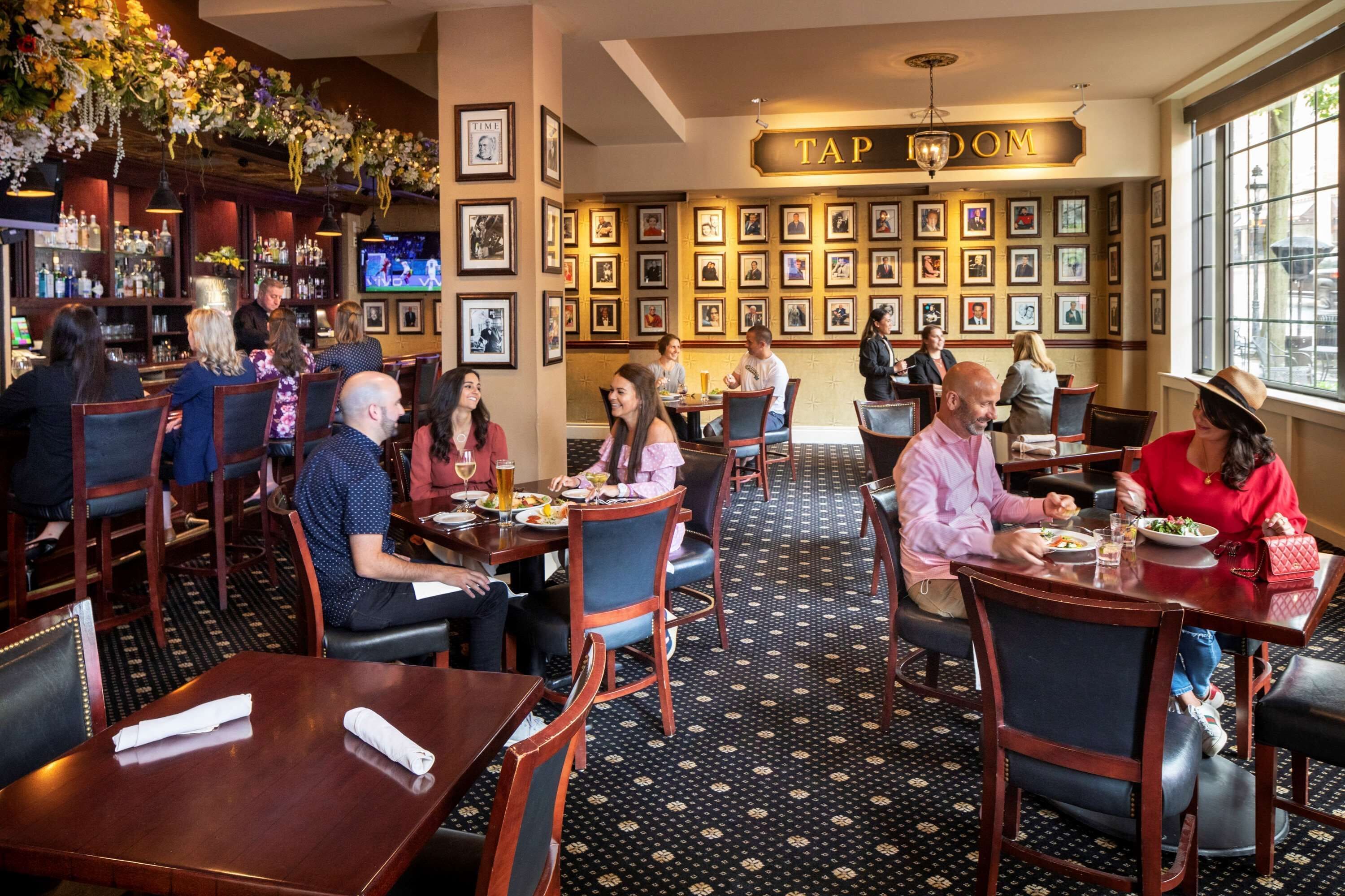 Restaurante com mesas e cadeiras de madeira, área de bar com garrafas e quadros nas paredes.