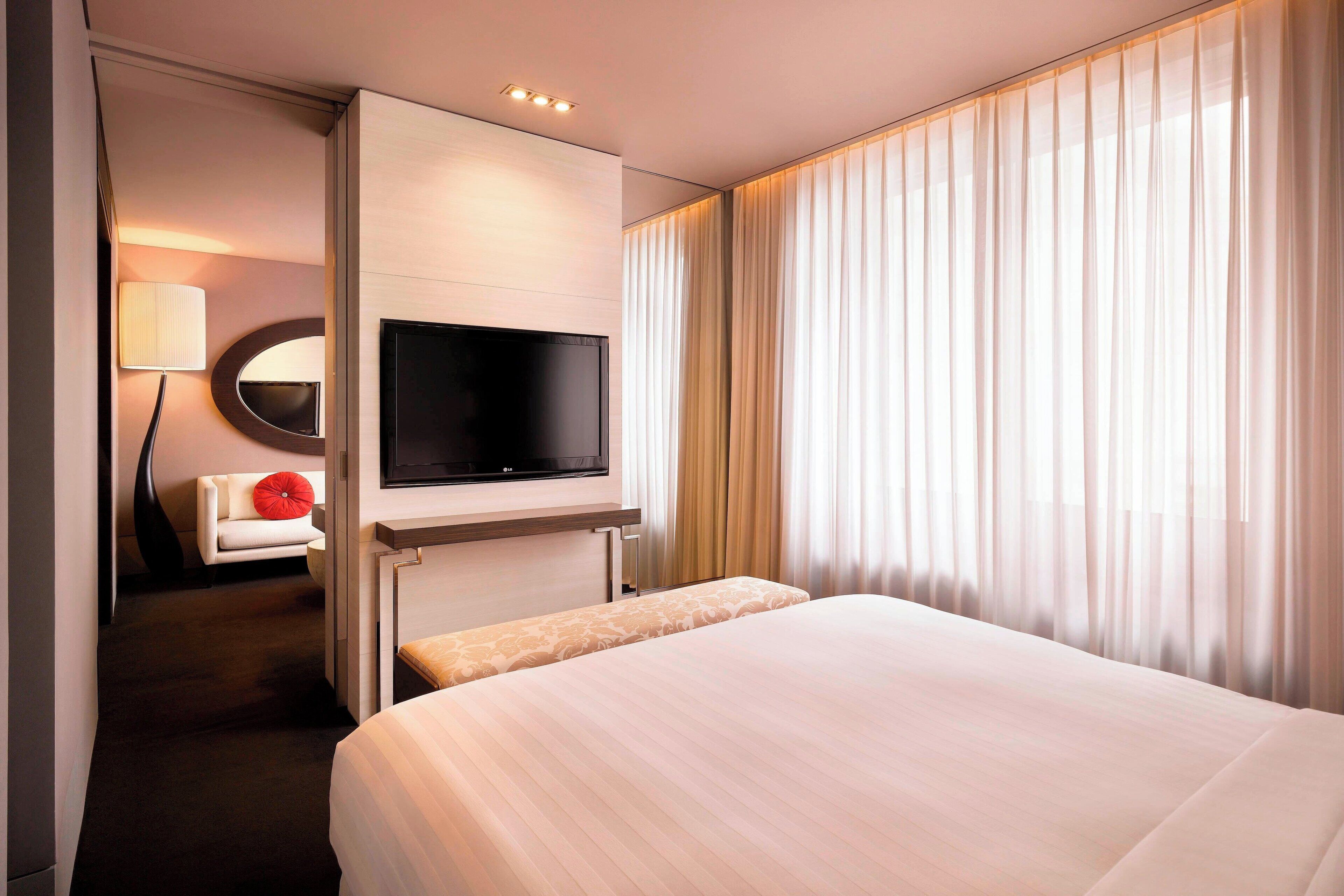 Una camera d'albergo con un letto, una TV a schermo piatto montata a parete, un divano con un cuscino rosso e un'ampia finestra con tende leggere.