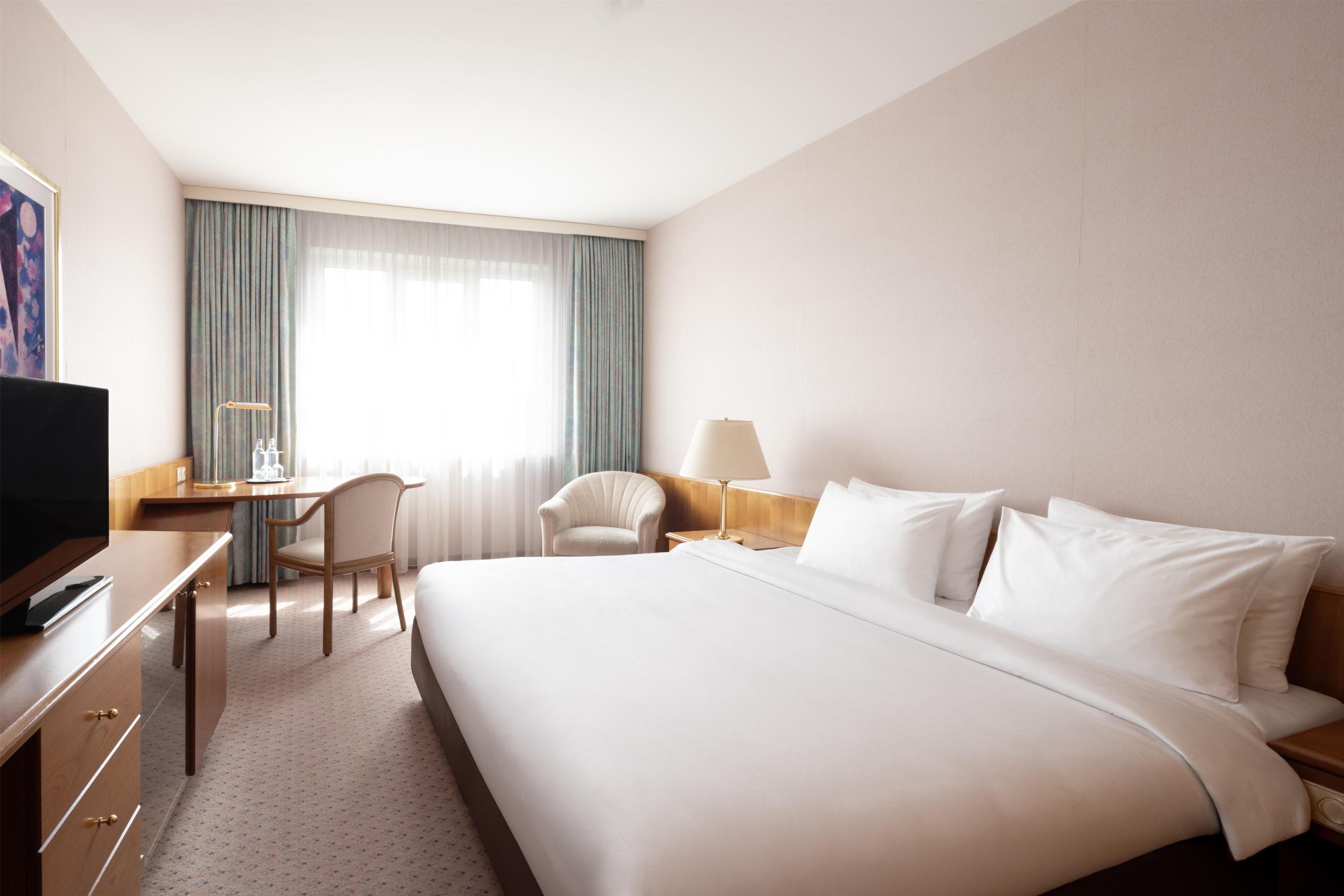 Una habitación de hotel con una cama grande, un escritorio con una silla, un televisor y una ventana con cortinas.