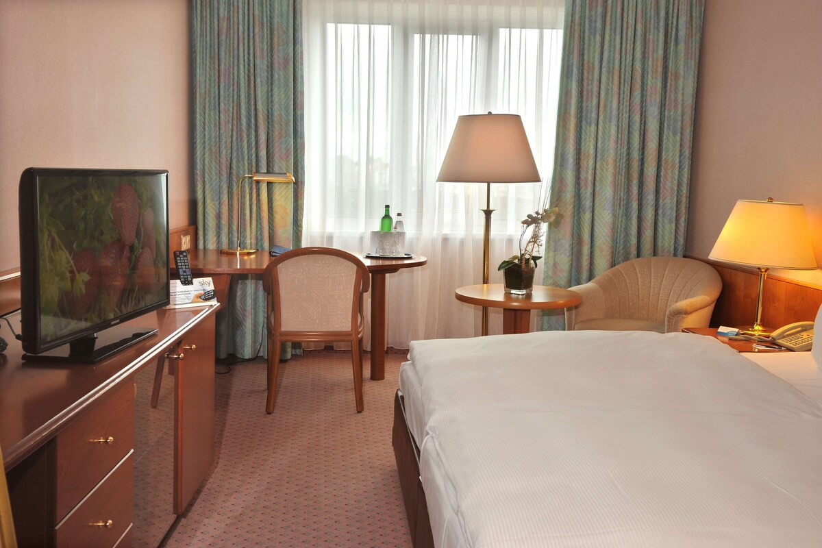 A hotel room with a bed, a desk with a television, a chair, a lamp, and a window with curtains.