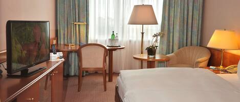 Quarto de hotel com cama, escrivaninha com televisão, cadeira, luminária e janela com cortinas.