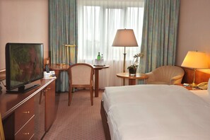 A hotel room with a bed, a desk with a television, a chair, a lamp, and a window with curtains.