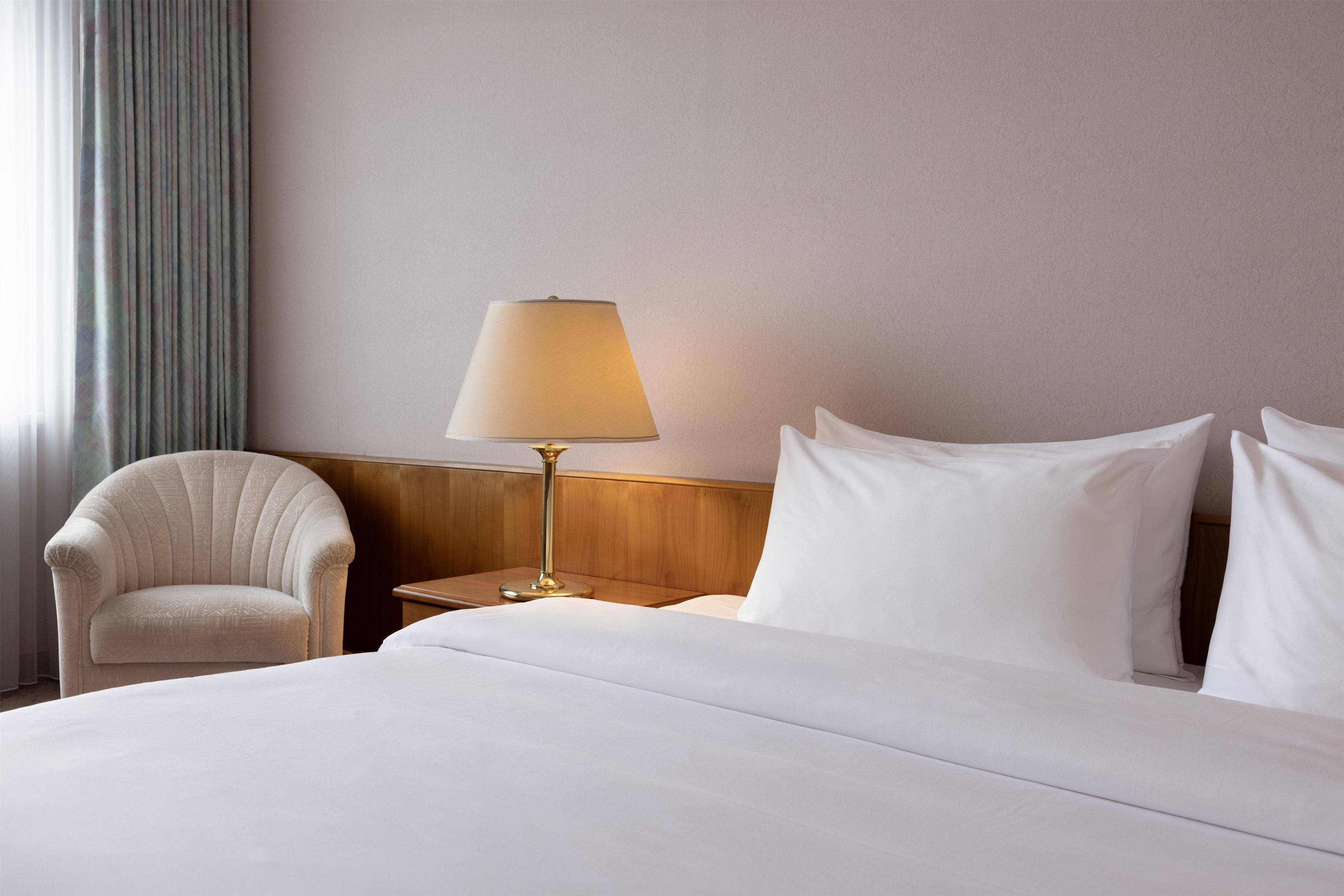A hotel room with a bed, a chair, a nightstand, a lamp, and a window with curtains.