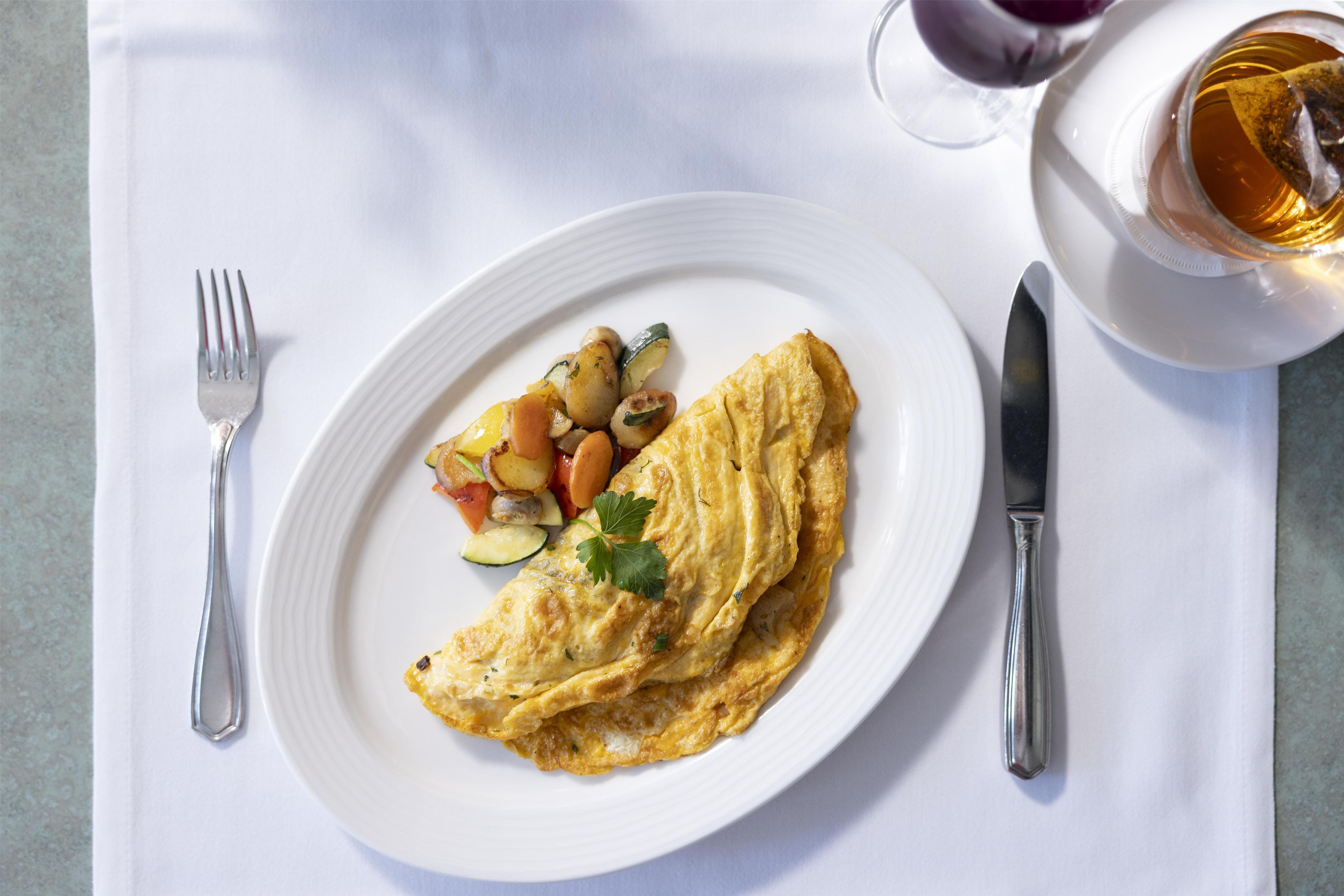A plate with an omelette and vegetables, a fork, a knife, and a cup of tea on a white tablecloth.