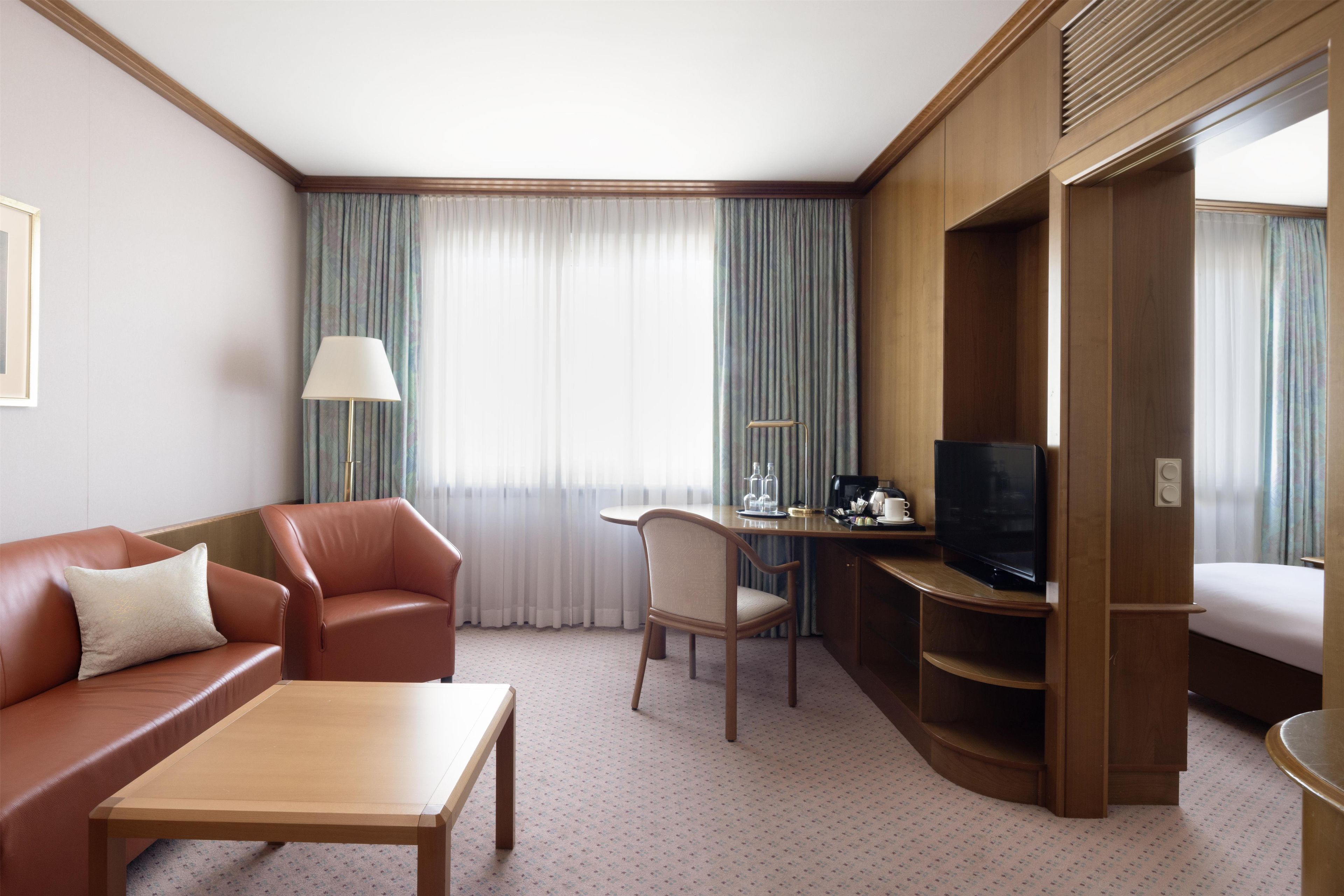 Ein Hotelzimmer mit Sofa, Schreibtisch, Fernseher, Sessel und Bett.