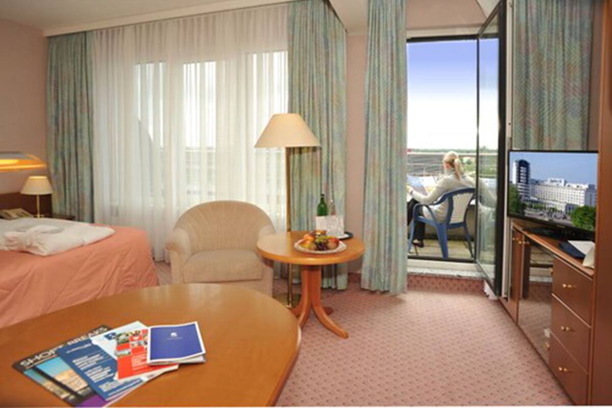 A hotel room with a bed, a desk with a lamp, a small table with a bottle and fruit, and a view of a cityscape through a glass door.