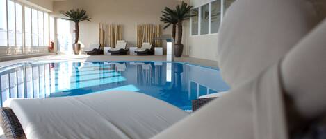 Una piscina interior moderna con agua cristalina azul, rodeada de sillones de descanso y palmeras.