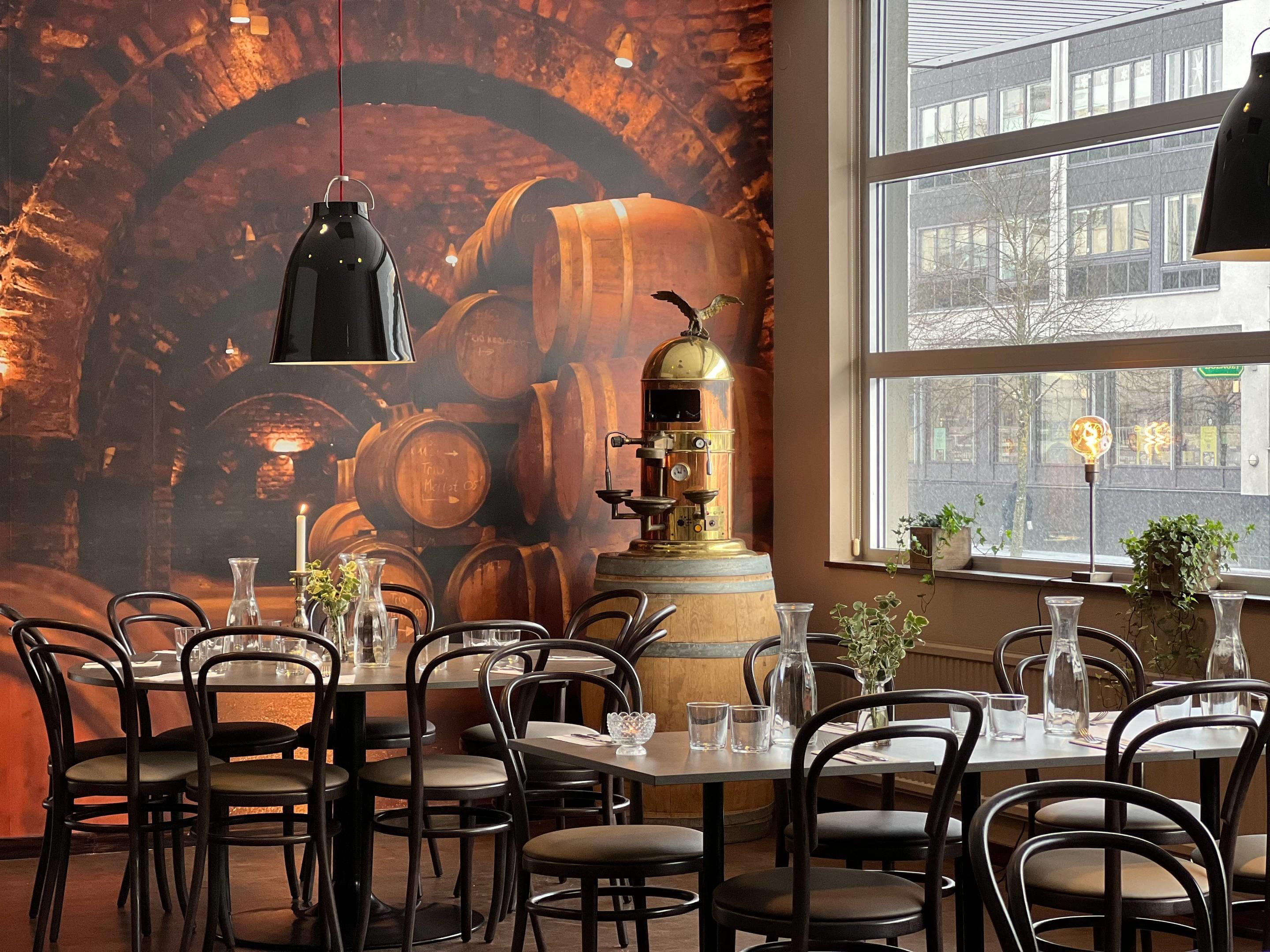 Ein Restaurantinneres mit einer Wandmalerei von Fässern, einer alten Espressomaschine und Blick auf die Stadt durch große Fenster.