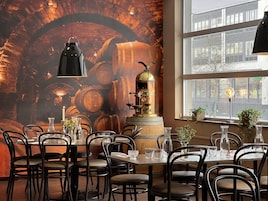 Interior de restaurante con un mural de barriles, una cafetera espresso antigua y vistas de la ciudad a través de amplias ventanas.