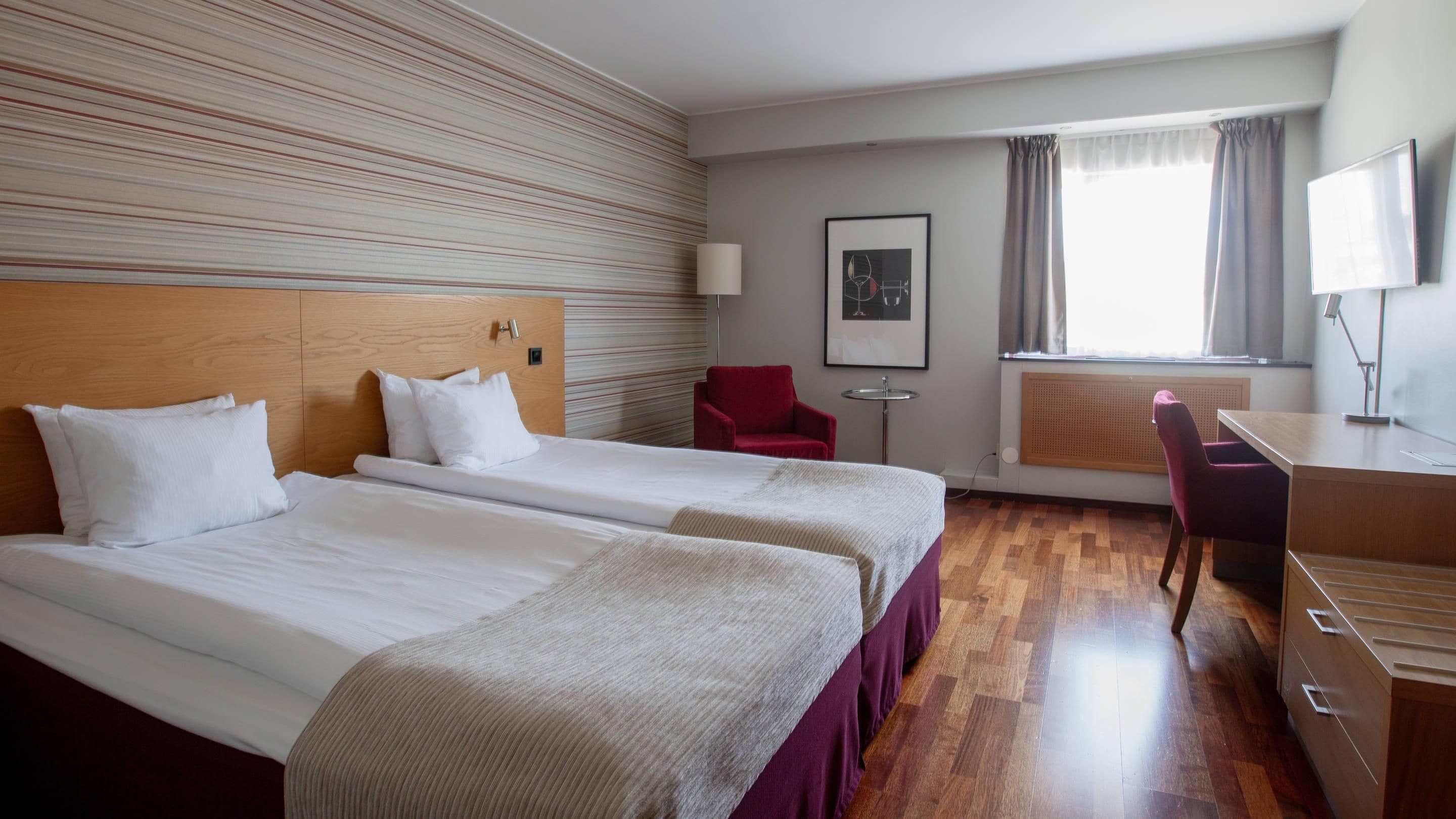Ett hotellrum med två sängar, ett skrivbord, en stol, en TV och ett fönster med gardiner.