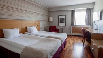 Ett hotellrum med två sängar, ett skrivbord, en stol, en TV och ett fönster med gardiner.