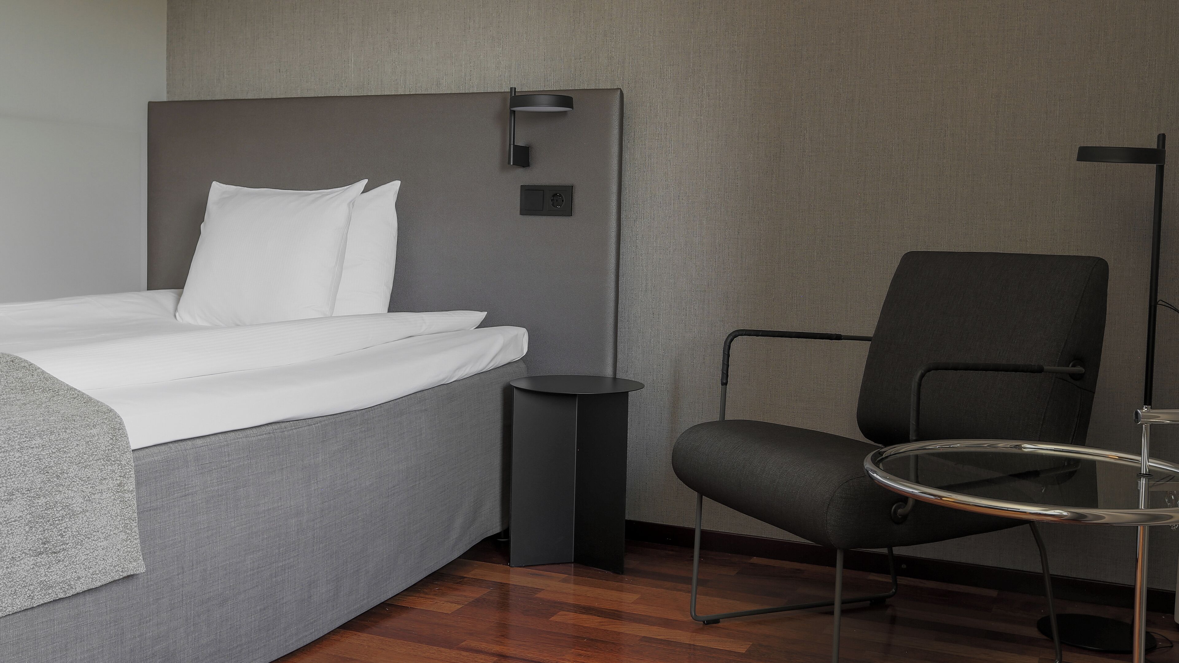 Et moderne hotelværelse med en pænt redt seng, en grå polstret stol, et lille rundt bord og en lampe monteret på væggen.