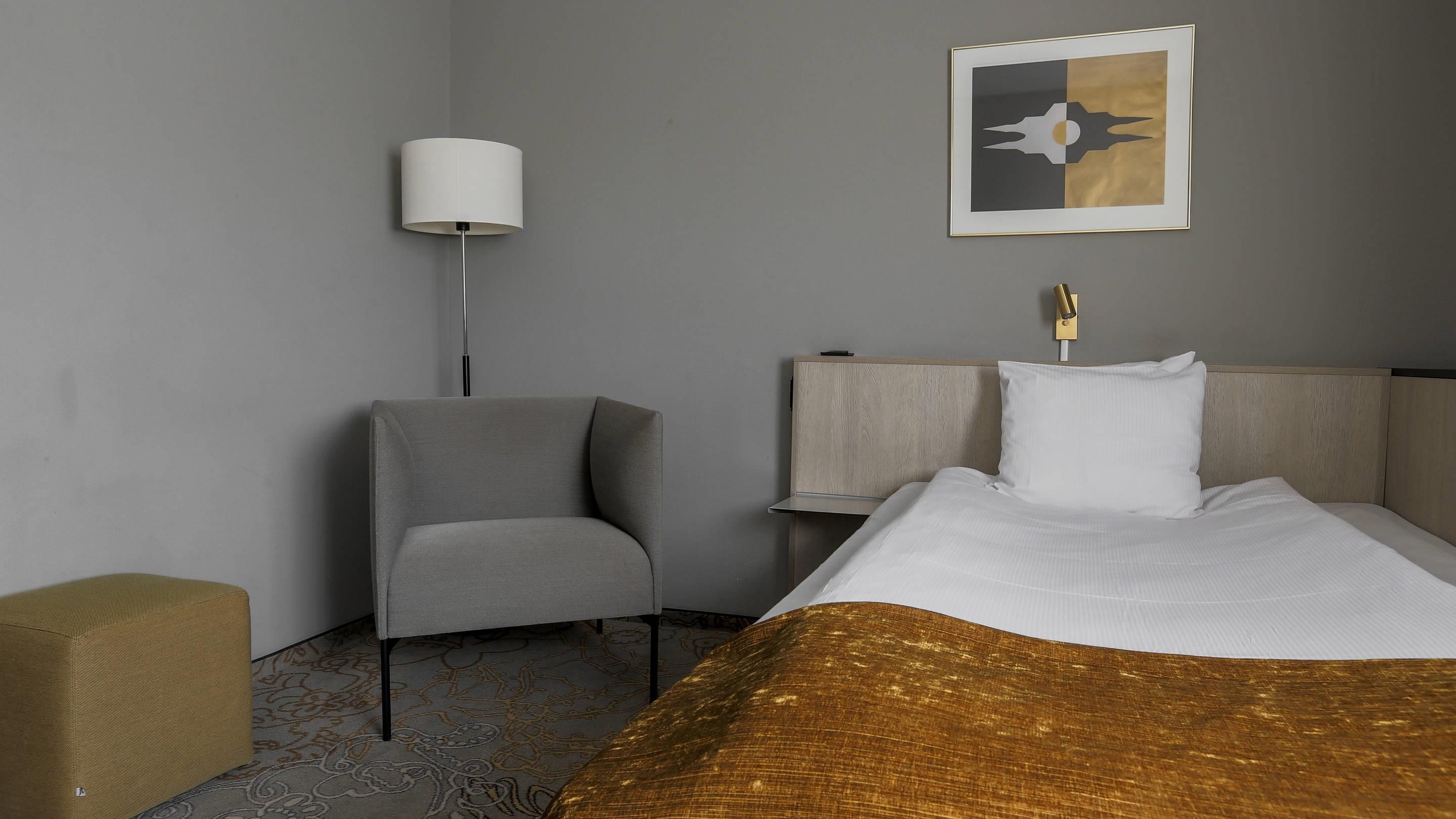 A hotel room with a bed, a chair, a bedside table, a lamp, and a wall art piece.