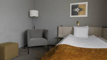 A hotel room with a bed, a chair, a bedside table, a lamp, and a wall art piece.