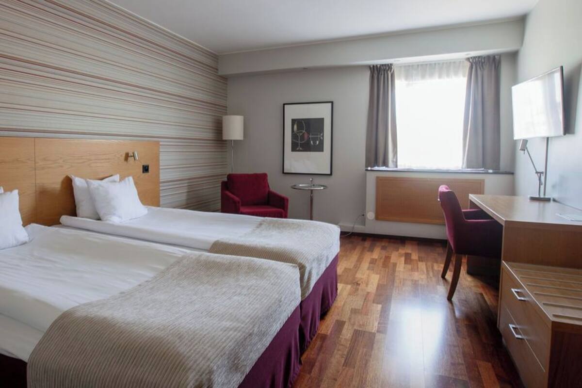 A hotel room with two beds, a desk, a chair, a lamp, and a window with curtains.