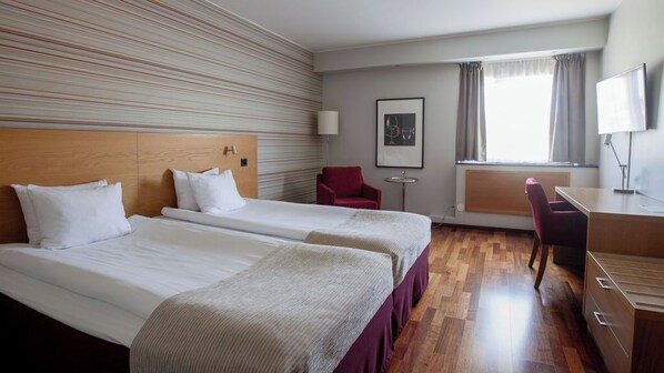 A hotel room with two beds, a desk, a chair, a lamp, and a window with curtains.