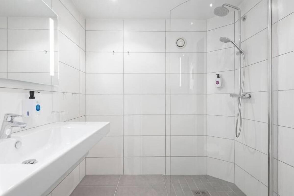 A modern bathroom with a white tiled shower, a sink with a mirror, and a soap dispenser.