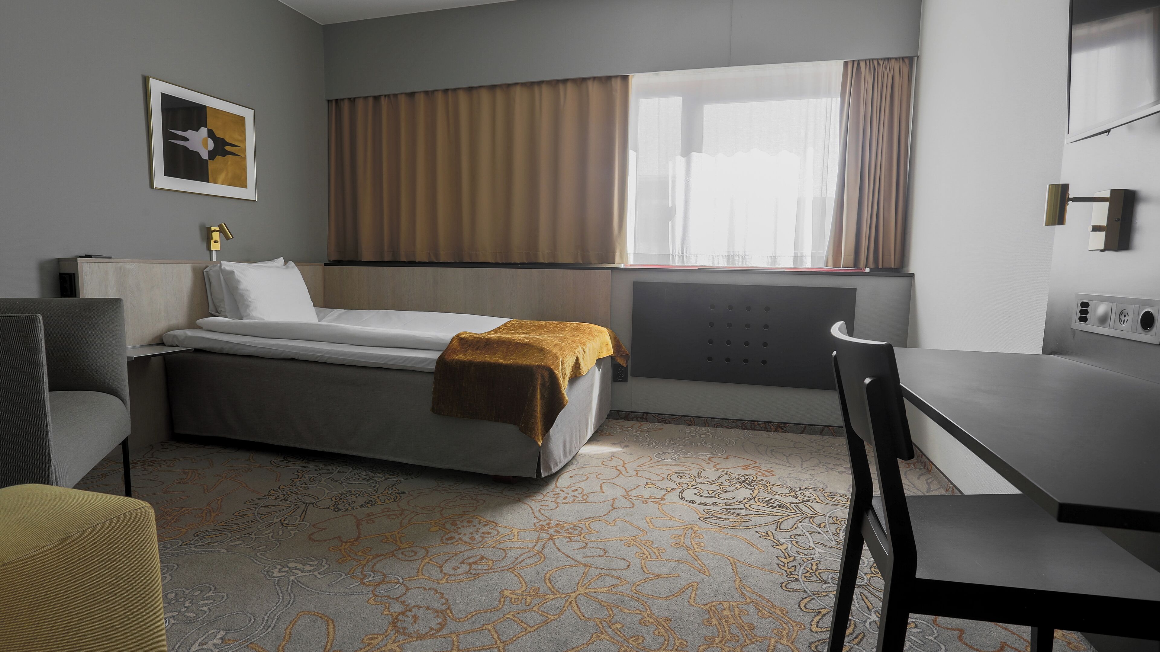 A hotel room with a bed, a desk, a chair, a window with curtains, a radiator, and a patterned carpet.