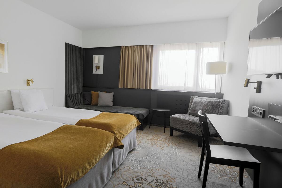 A hotel room with two beds, a desk, a chair, a sofa, and a window with curtains.